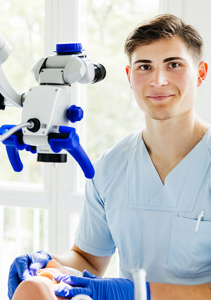 Young smiling dental professional in light-blue scrubs and blue gloves working on a dental mannequin with instruments beneath a white-and-blue dental microscope in a bright clinic setting.