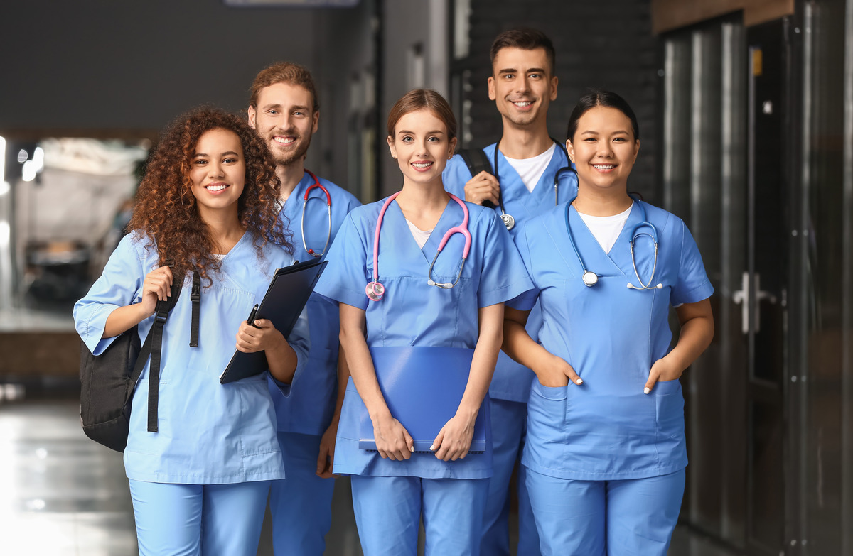 Five smiling healthcare workers in blue scrubs stand together in a hospital corridor, several wearing stethoscopes and one holding a clipboard while another carries a backpack.
