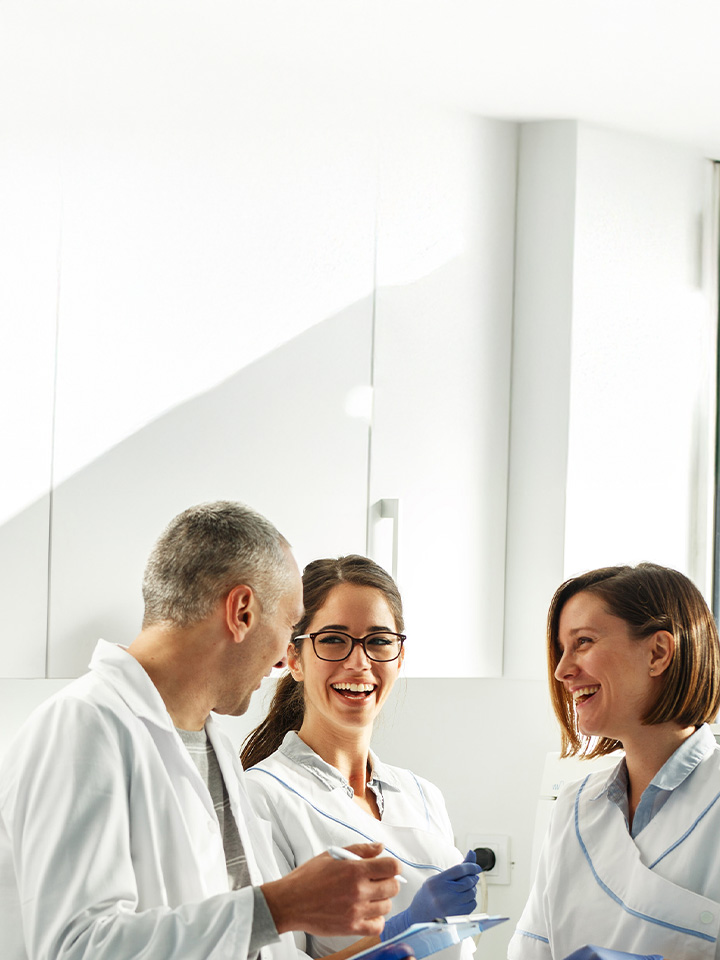 Three healthcare professionals in white coats stand in a bright clinical room, smiling and talking while one holds a clipboard and another wears blue examination gloves.