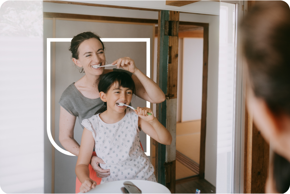 A mother stands behind her smiling young daughter as they brush their teeth together at a bathroom sink, both looking into the mirror.
