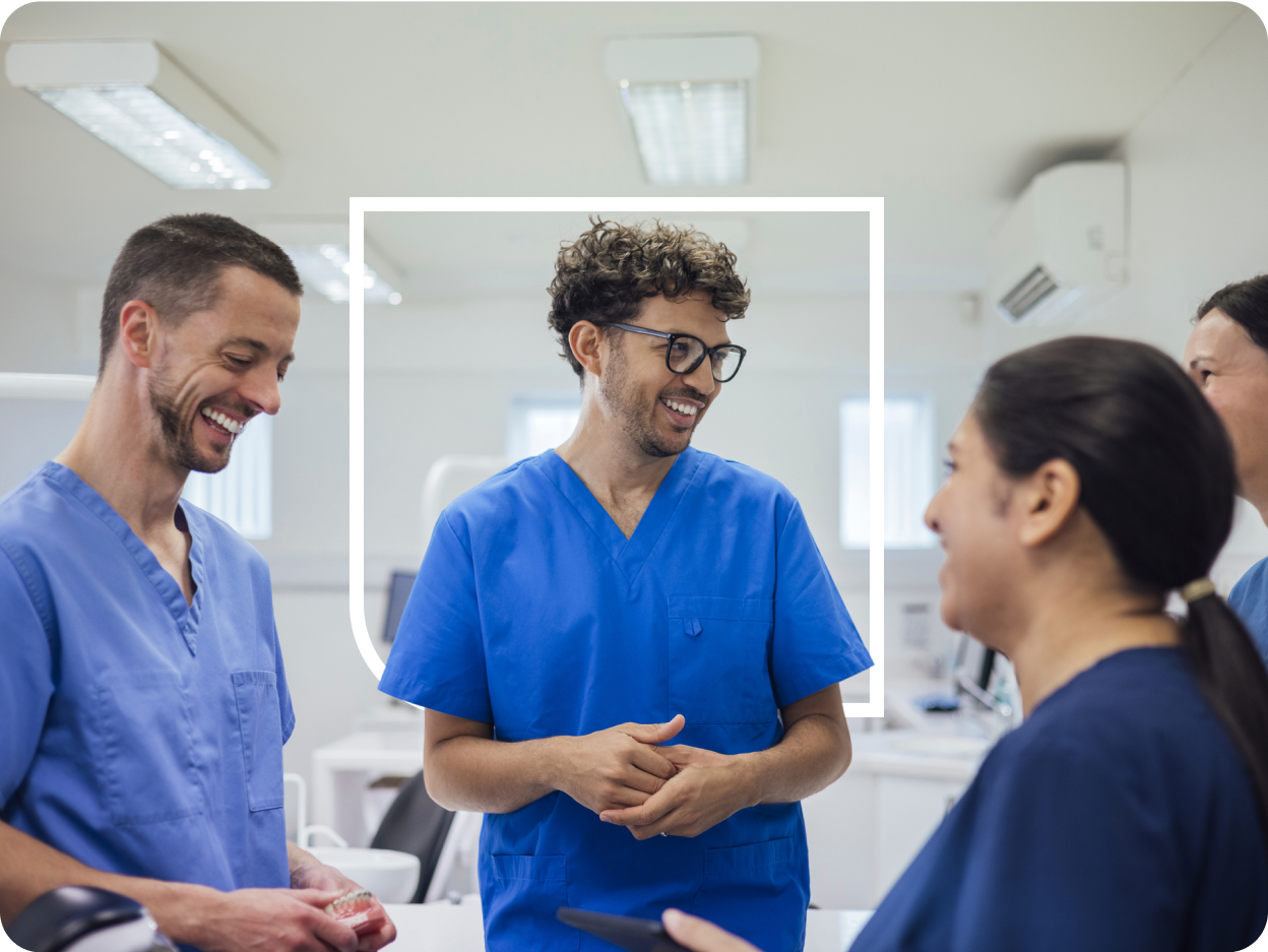 Four healthcare workers in blue scrubs standing in a bright clinic, smiling and chatting as the central man with curly hair and glasses speaks while colleagues listen, one holding a dental model and another holding a tablet.