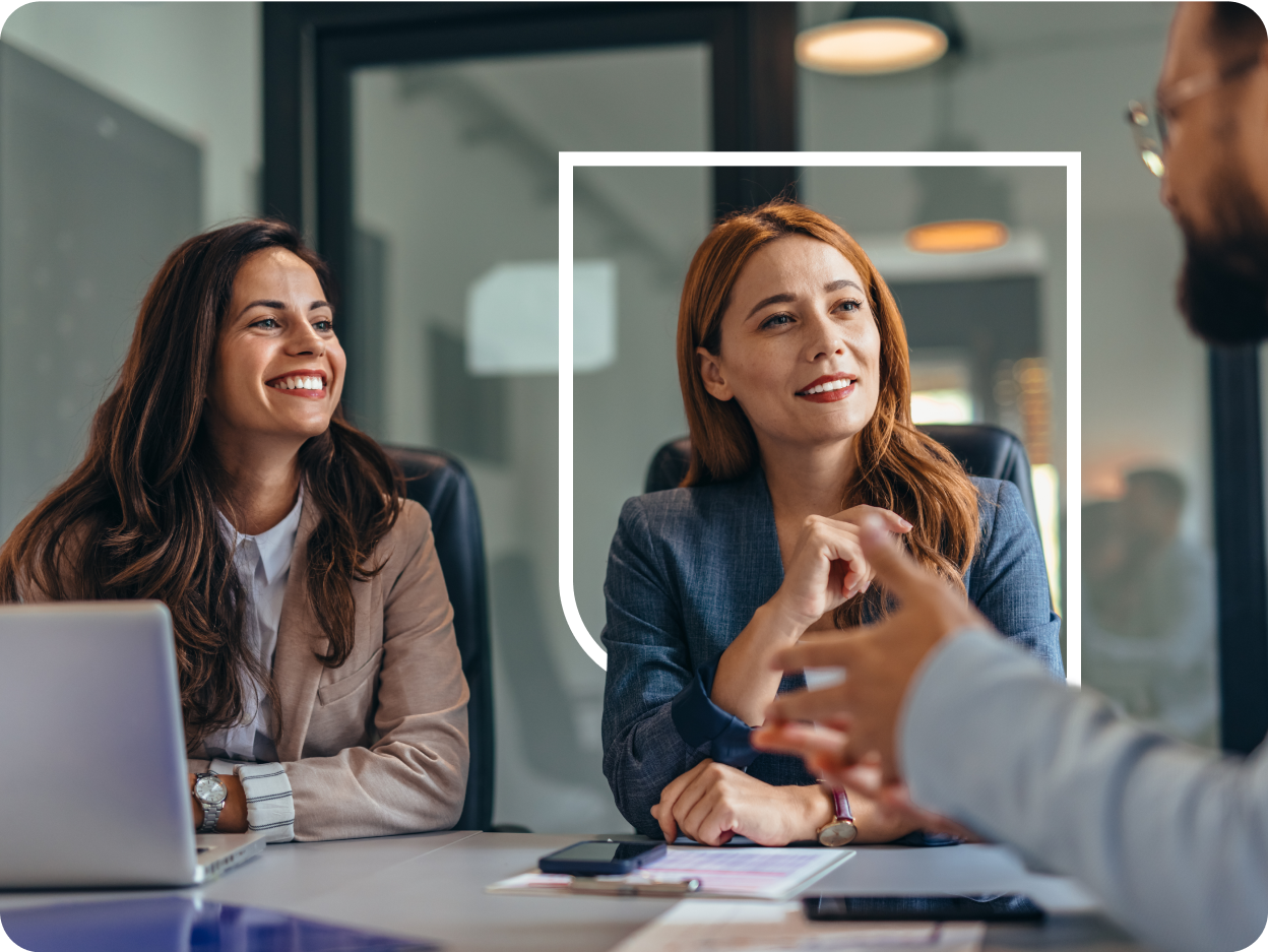Two professional women seated at a conference table—one in a tan blazer smiling broadly and the other in a blue blazer attentively listening while a colleague’s blurred hands gesture in the foreground, with a laptop, documents, and an office interior visible.