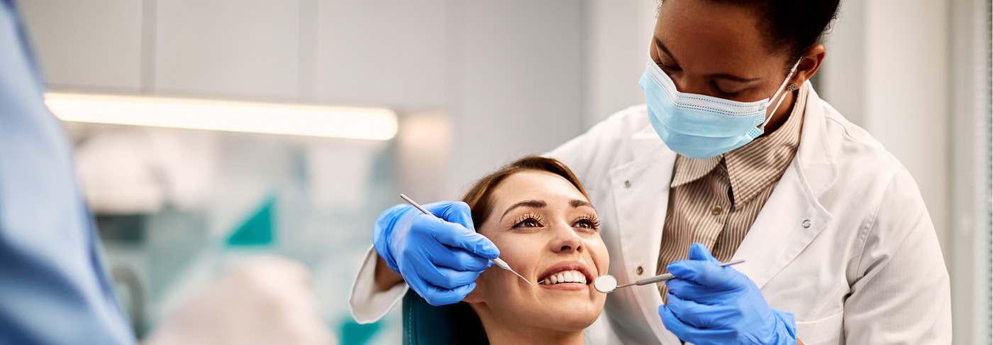 Dentist wearing a surgical mask and blue gloves examines a smiling patient's teeth with a dental mirror and probe while the patient reclines in a bright clinic.