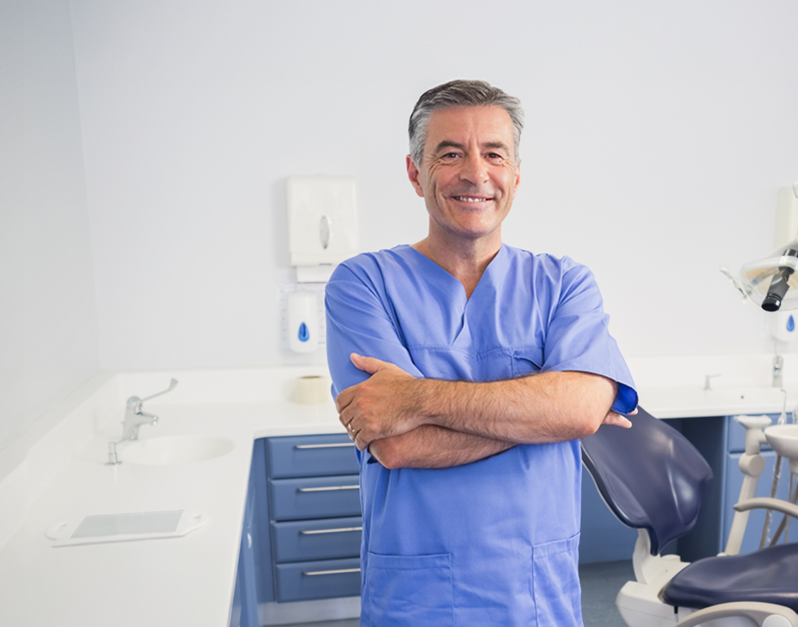 Smiling middle-aged male dentist with short gray hair wearing blue scrubs, standing with arms crossed beside a dental chair and cabinets in a bright dental exam room.