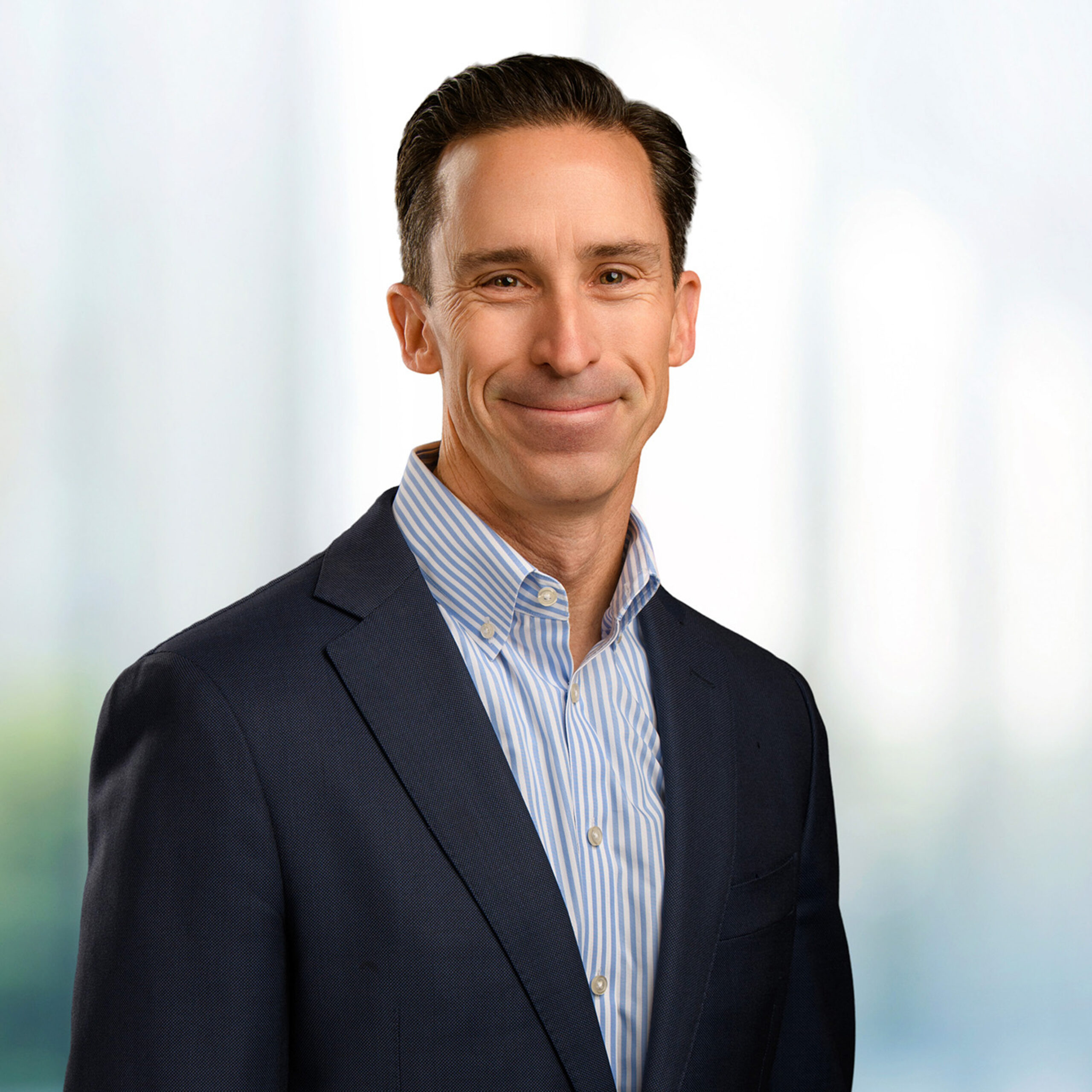 Middle-aged man with short dark hair wearing a navy suit and blue-striped shirt, smiling against a soft blurred background
