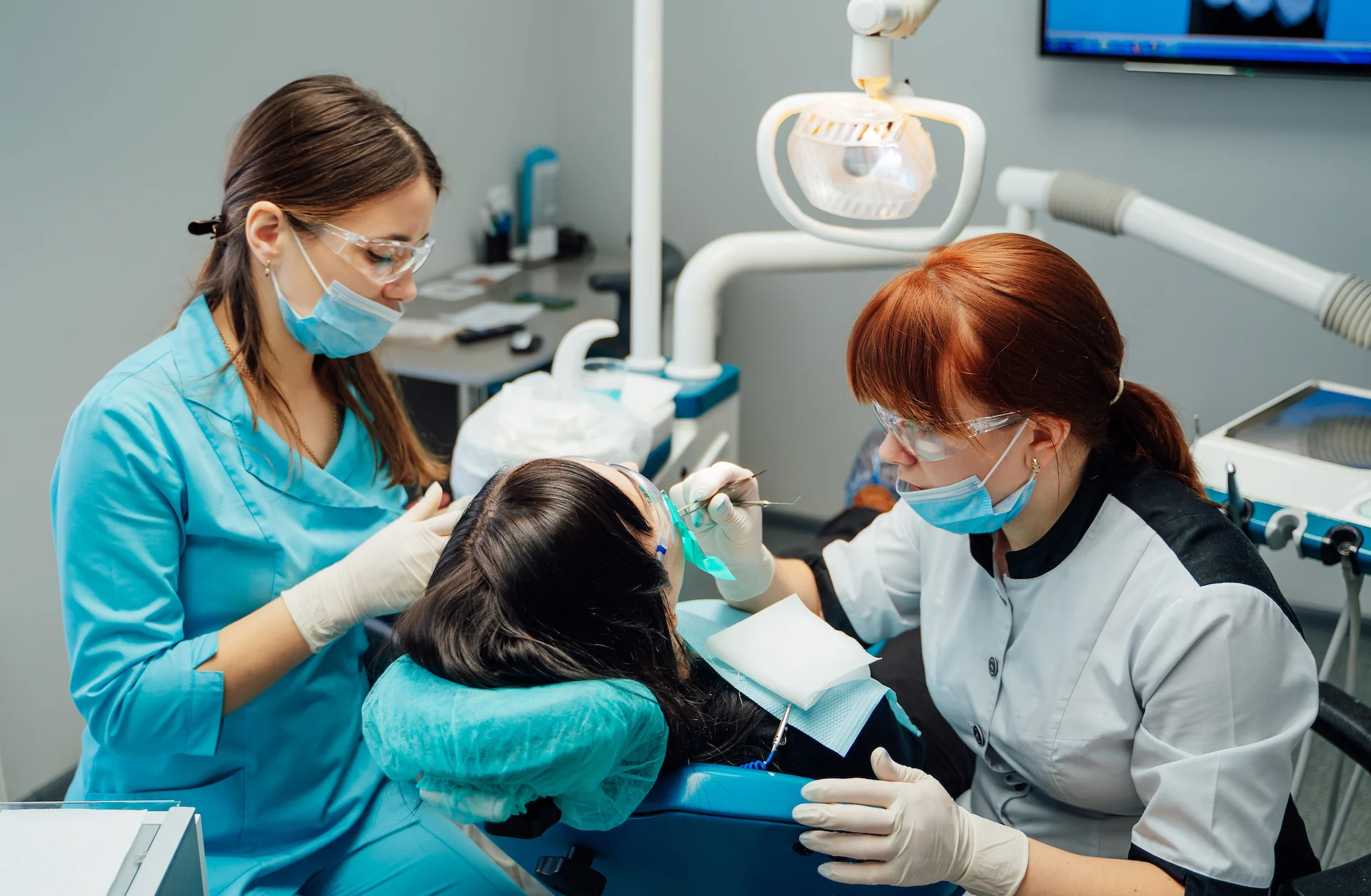 Two masked dental clinicians in gloves and protective goggles examine a patient reclined in a dental chair under an overhead lamp, one holding dental instruments while the other assists.