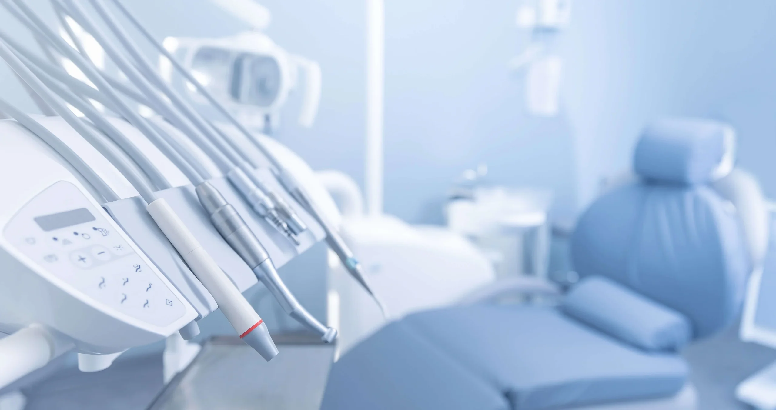 Close-up of dental handpieces and a control panel on a treatment unit in sharp focus, with a blurred light-blue dental chair and clinic equipment in the background.