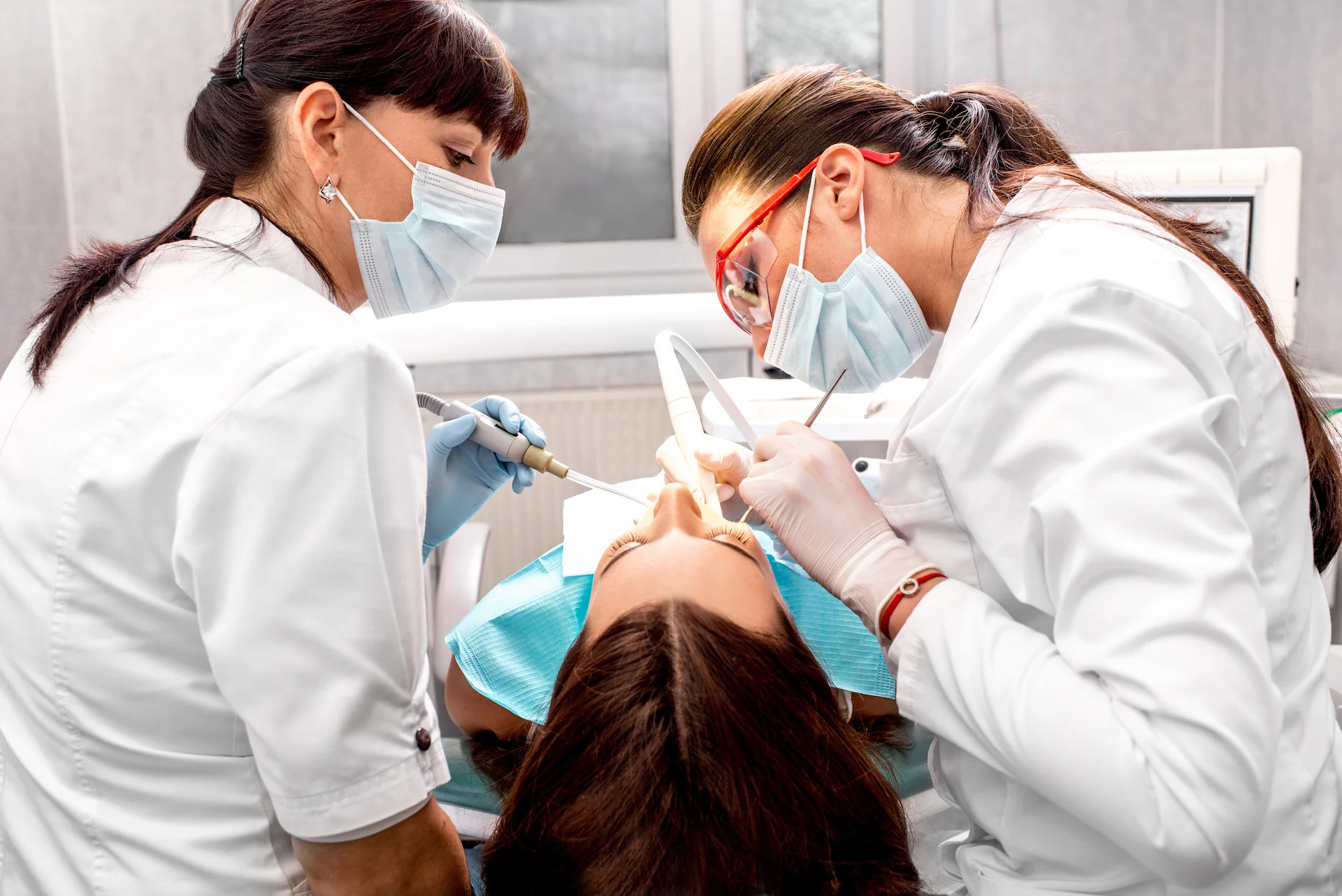 Two masked dental professionals in white coats lean over a reclining patient wearing a blue bib, using dental instruments and suction while working in a bright clinic setting.