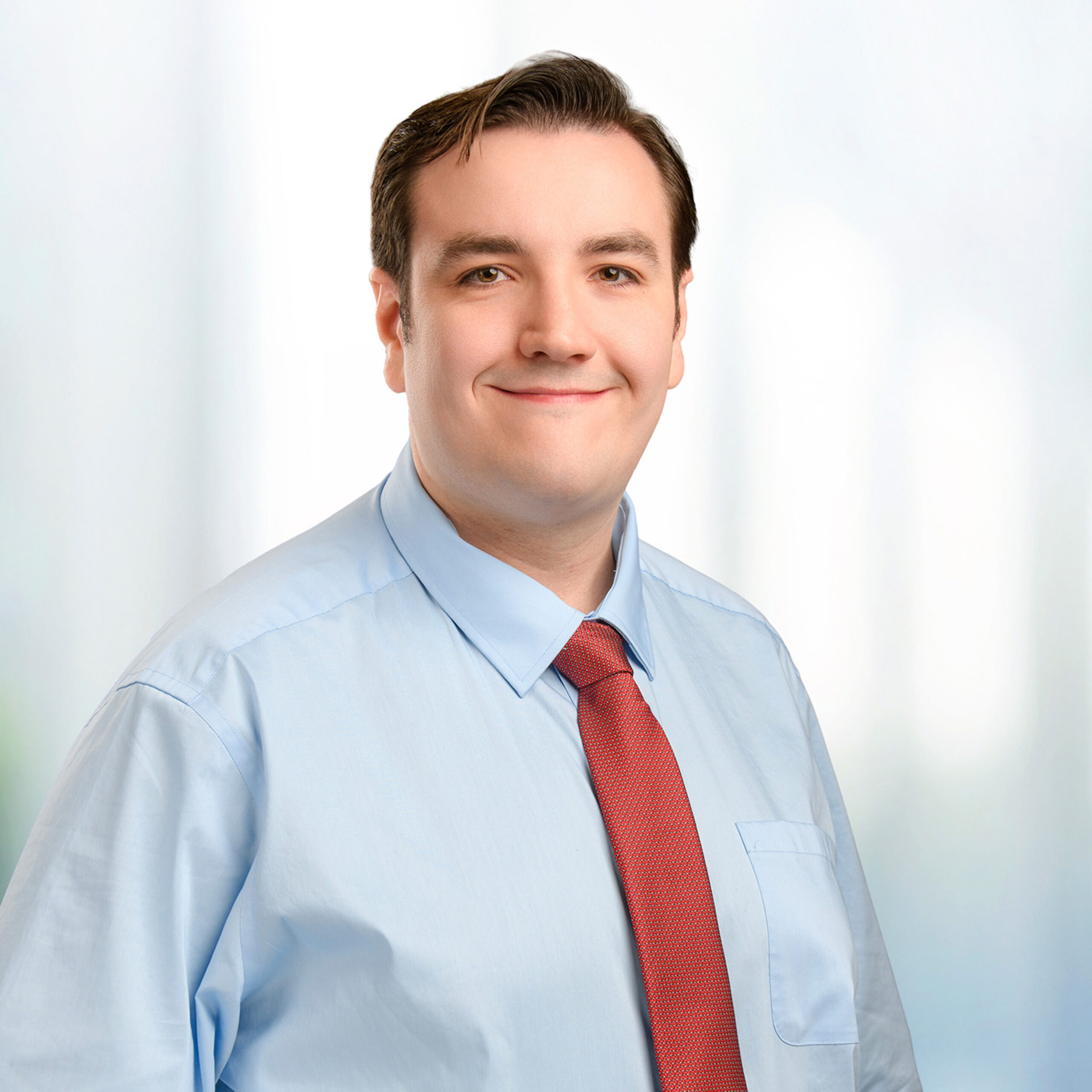 Young man in a light blue dress shirt and red tie smiling at the camera against a soft, blurred background.