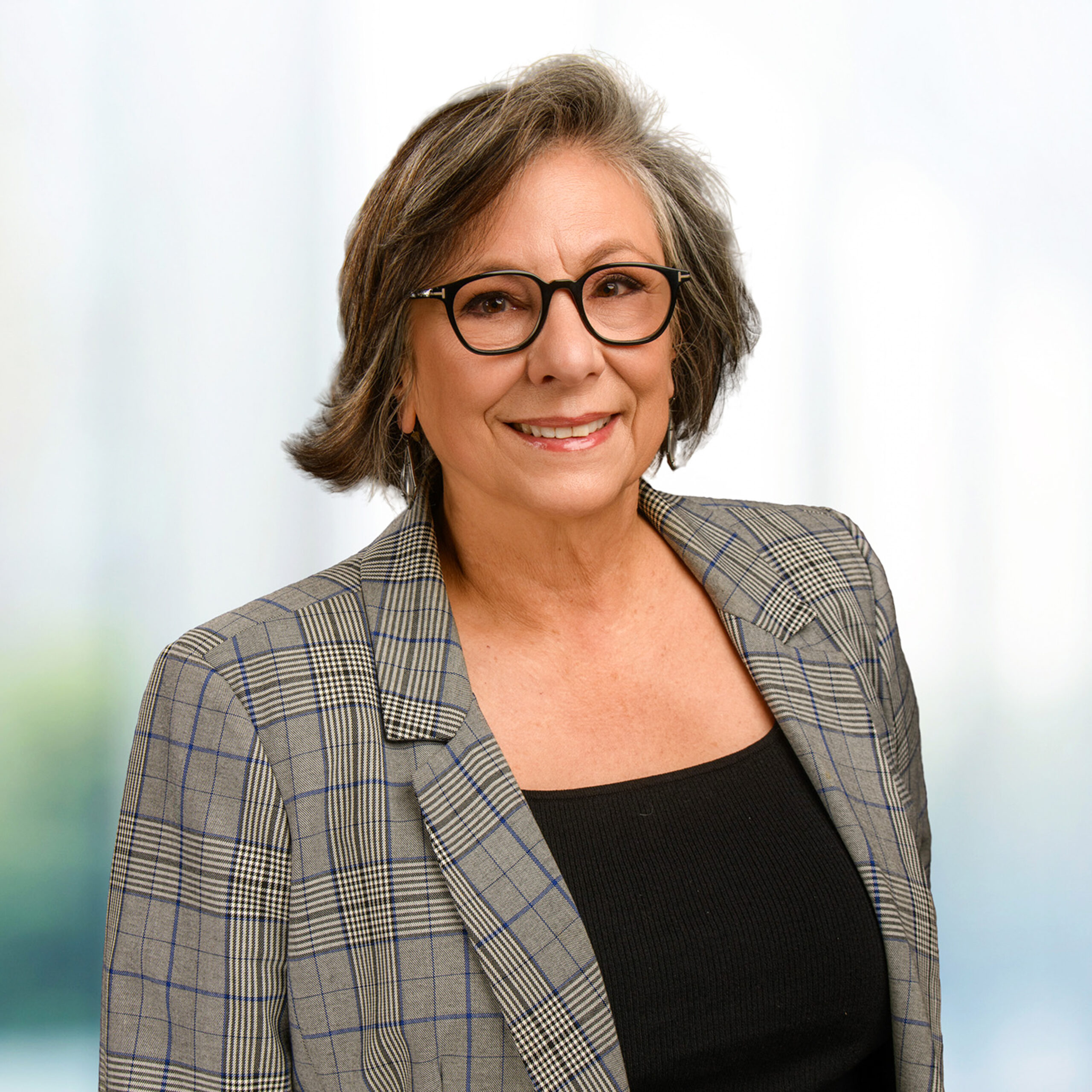Smiling woman with short gray hair wearing round black glasses and a gray plaid blazer over a black top.