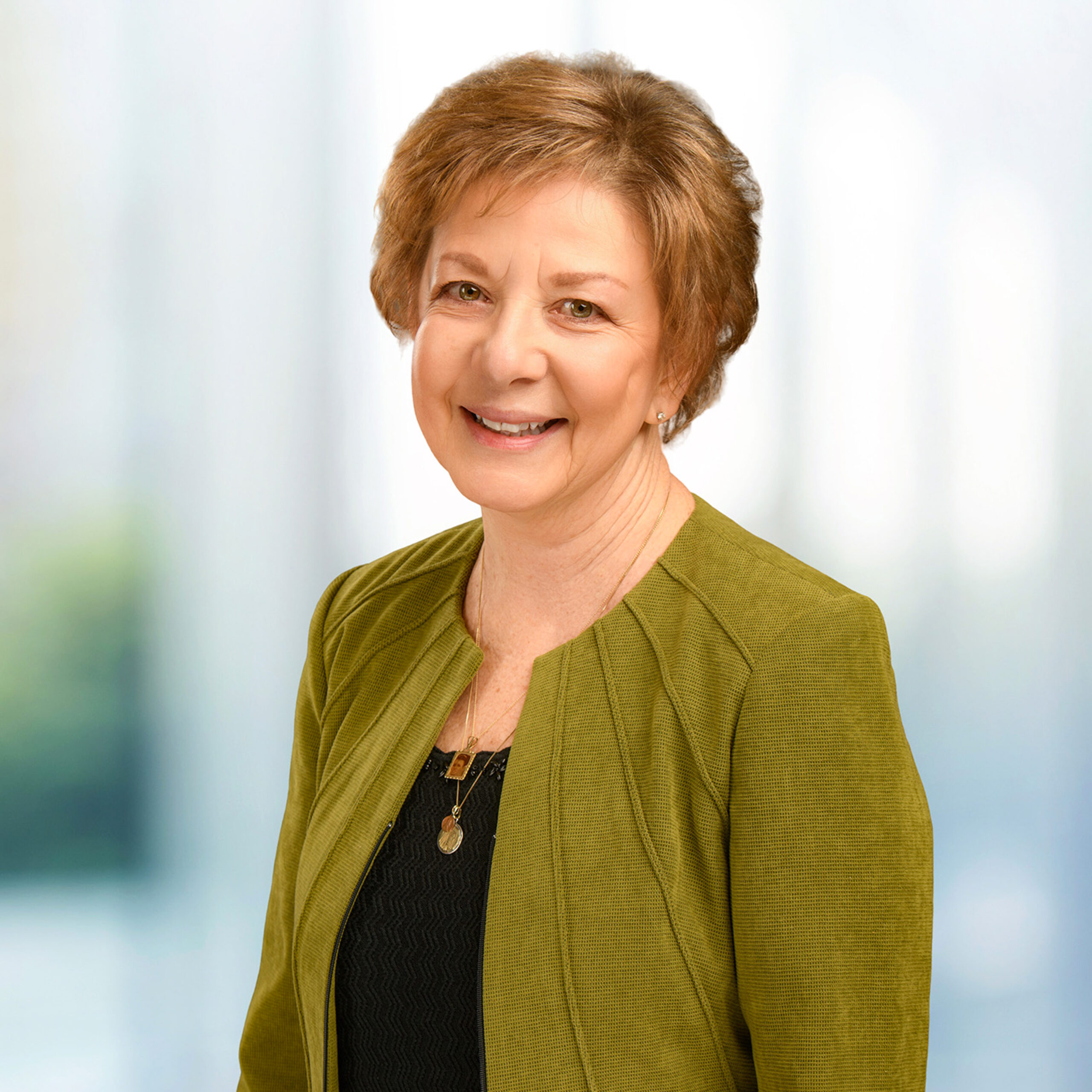 Smiling older woman with short light brown hair wearing a green jacket and pendant necklaces against a blurred background.