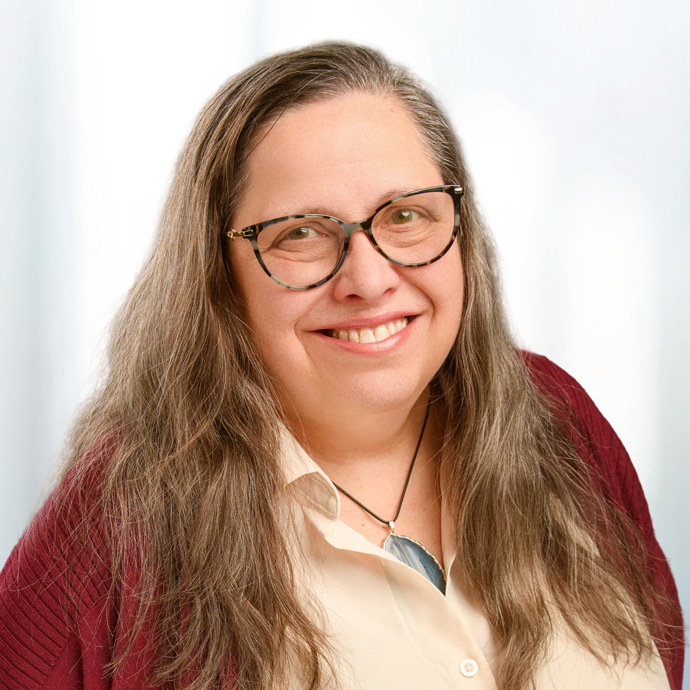 Smiling woman with long gray hair and tortoiseshell glasses wearing a white blouse, maroon cardigan, and a blue stone pendant.