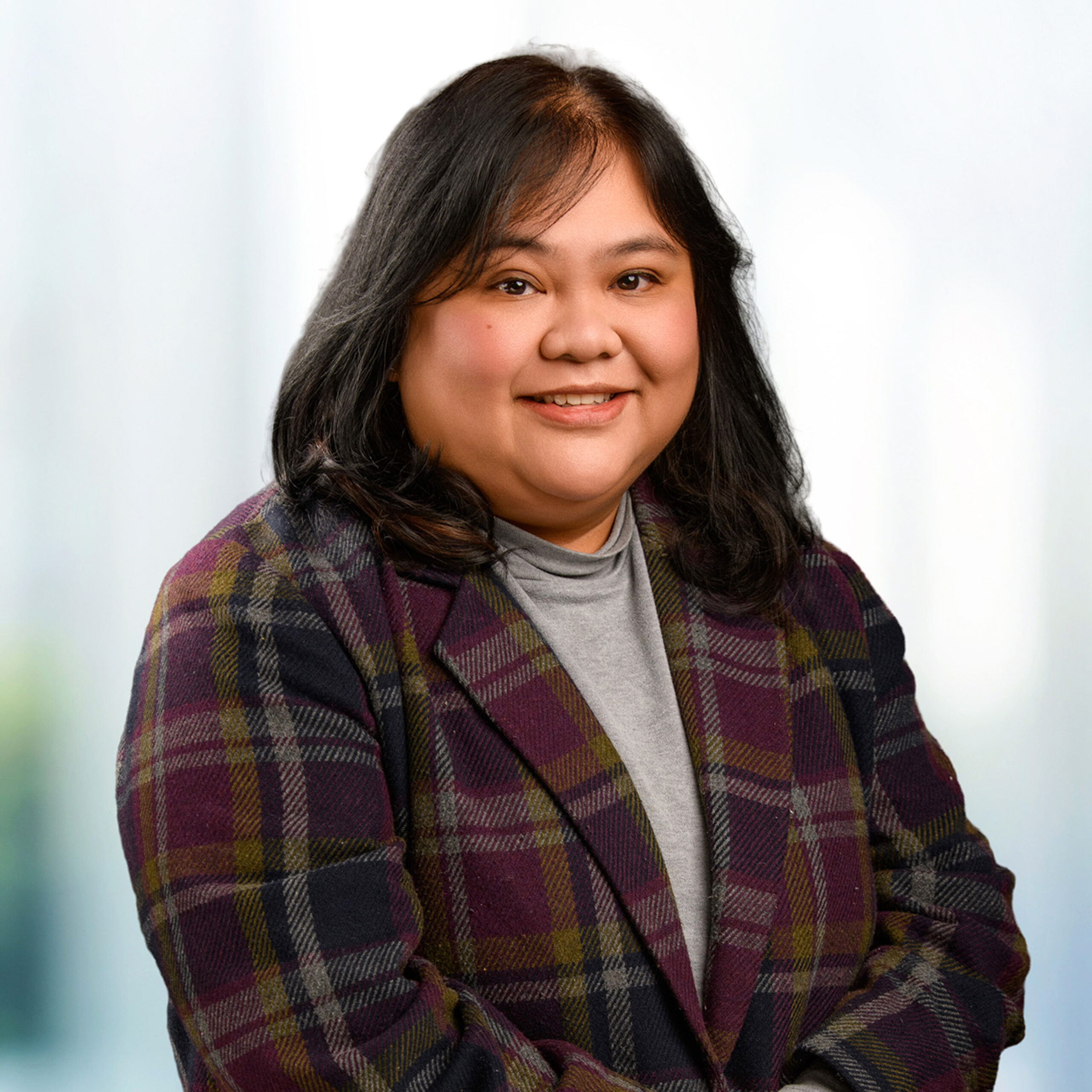 Smiling woman with shoulder-length dark hair wearing a gray turtleneck and purple plaid blazer against a soft blurred background.