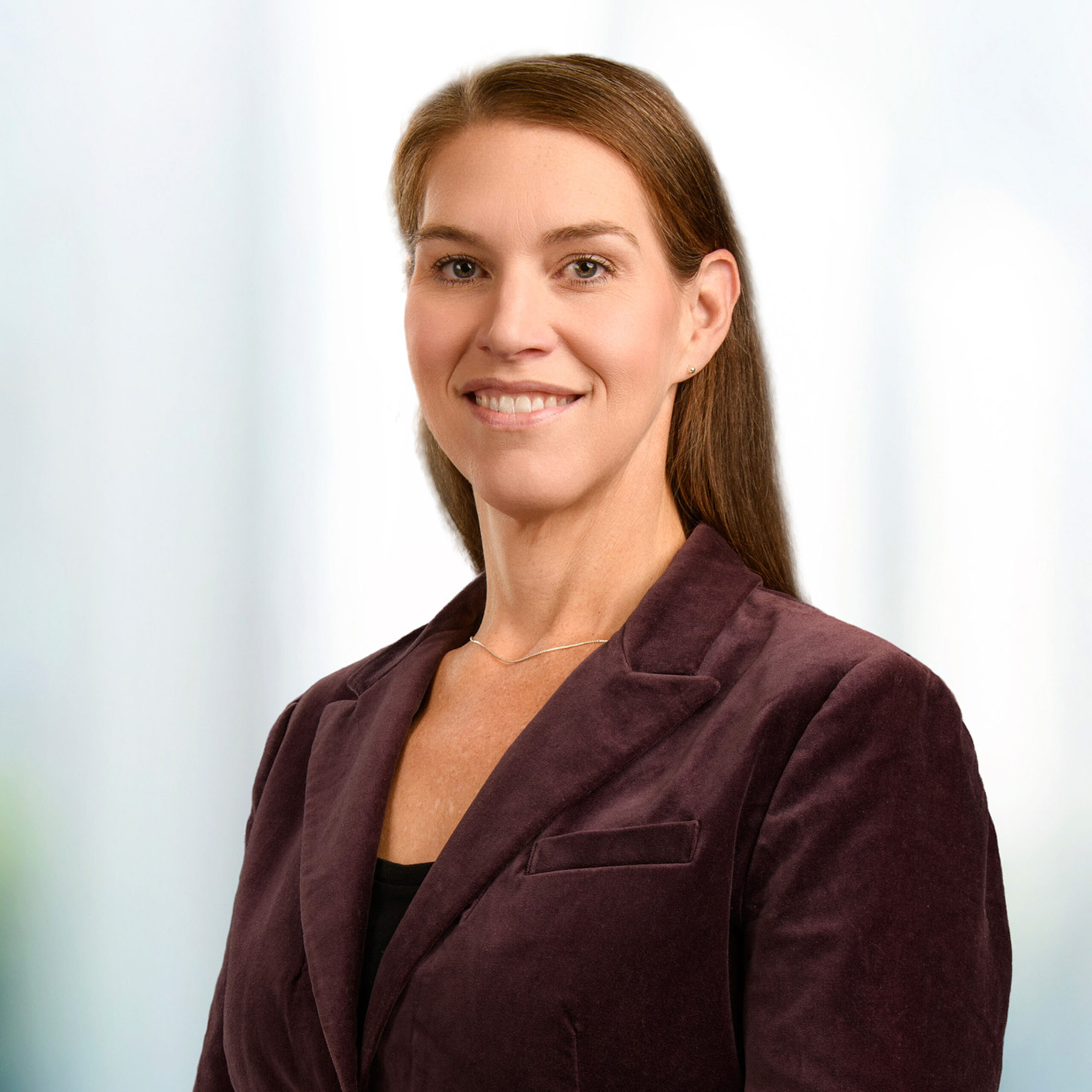Woman with brown hair in a plum blazer and thin necklace, smiling in a professional headshot against a soft background.