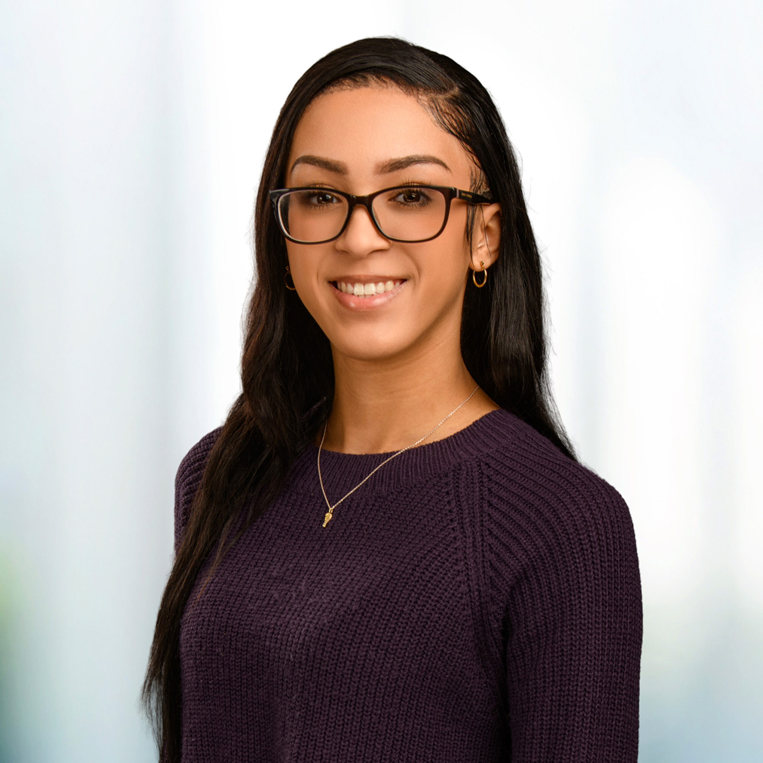 Smiling woman with glasses and long dark hair wearing a purple sweater and hoop earrings against a blurred background.