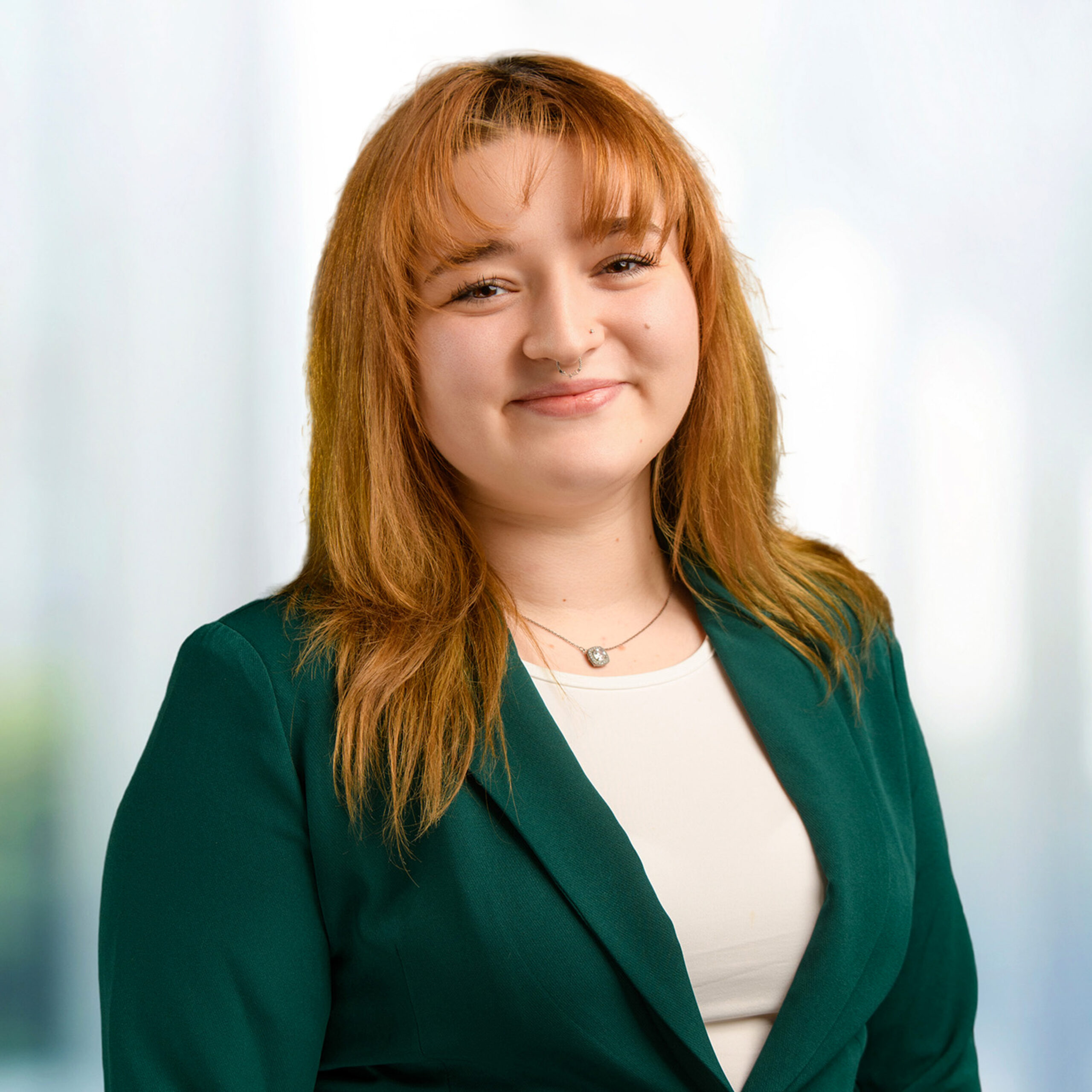 Young woman with auburn bangs and septum piercing, smiling in a green blazer over a white top against a blurred background.