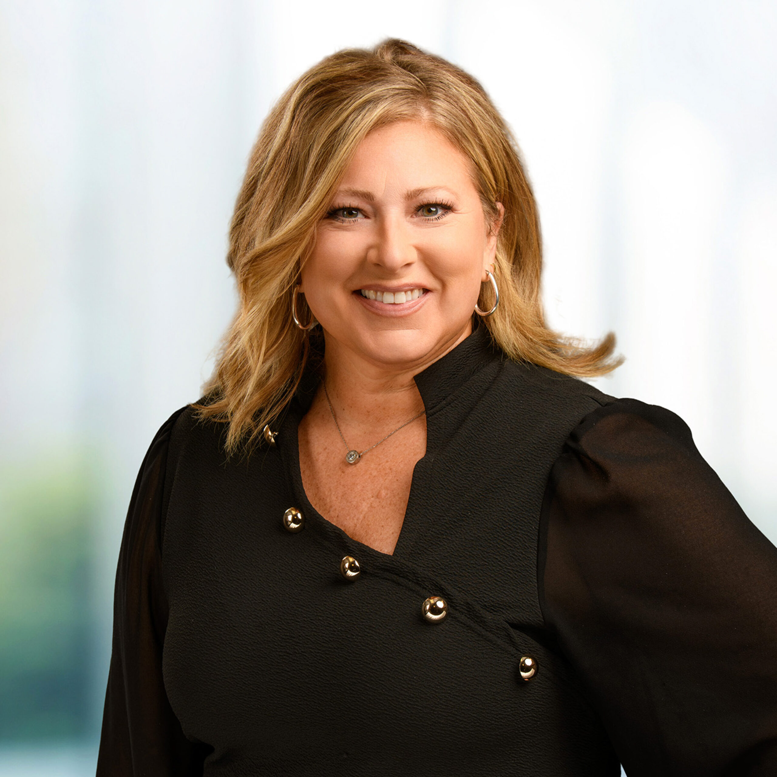 Smiling woman with shoulder-length blonde hair wearing a black blouse with gold buttons and hoop earrings against a soft, blurred background