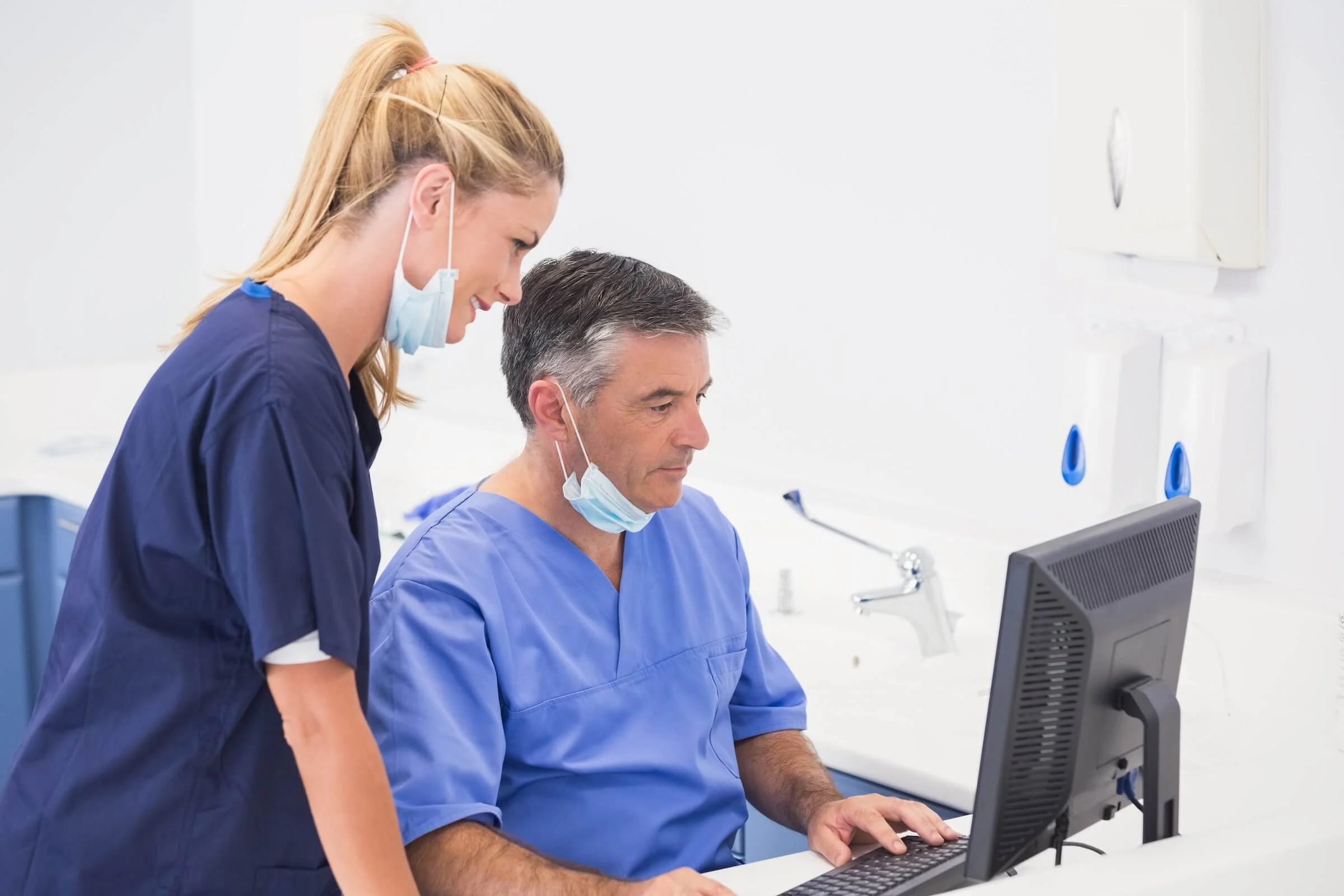 A male clinician in light blue scrubs sits at a computer typing while a female nurse in dark blue scrubs leans over to look at the monitor, both wearing surgical masks pulled below their chins in a bright clinical workspace.