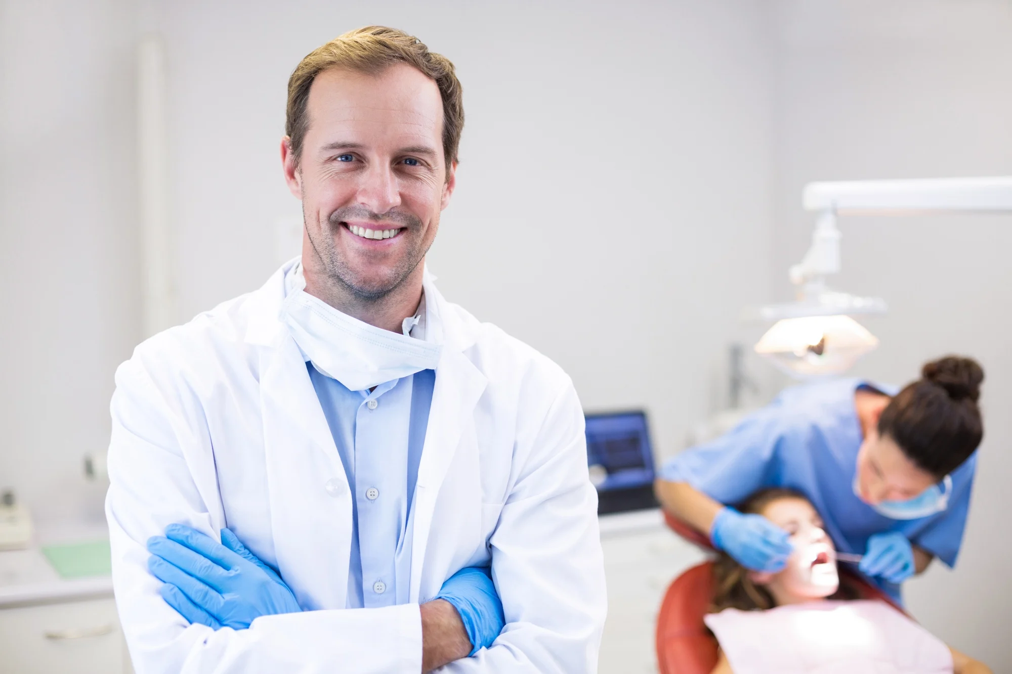 Smiling male dentist in a white coat with a mask around his neck and blue gloves stands with arms crossed in a dental clinic while a dental assistant treats a patient reclined under a lamp in the background.