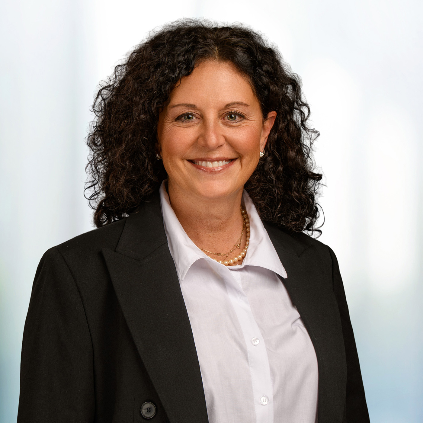 Smiling woman with dark curly hair wearing a black blazer, white blouse and pearl necklace against a soft blurred background.