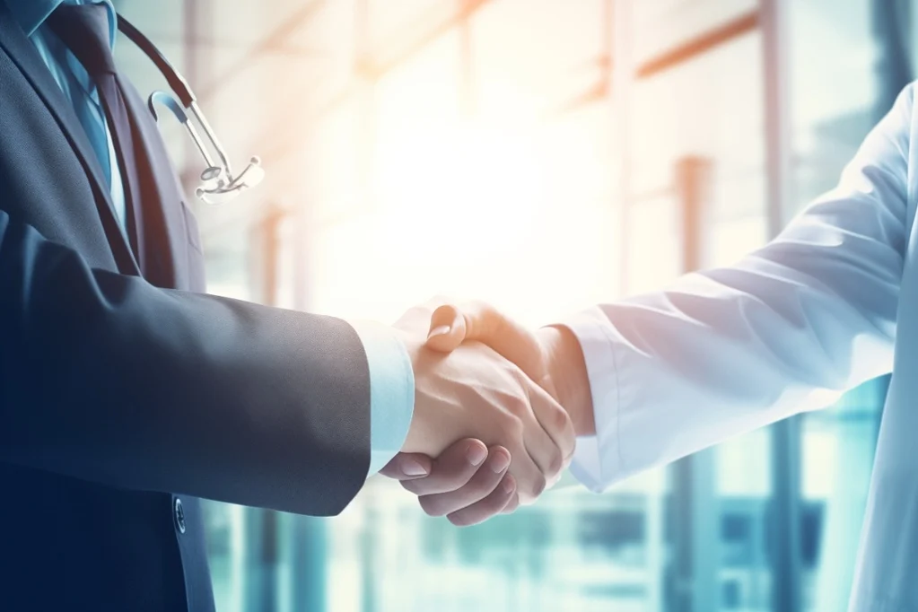 Businessperson in a suit and a doctor wearing a white coat and stethoscope shaking hands in a bright, modern medical office.