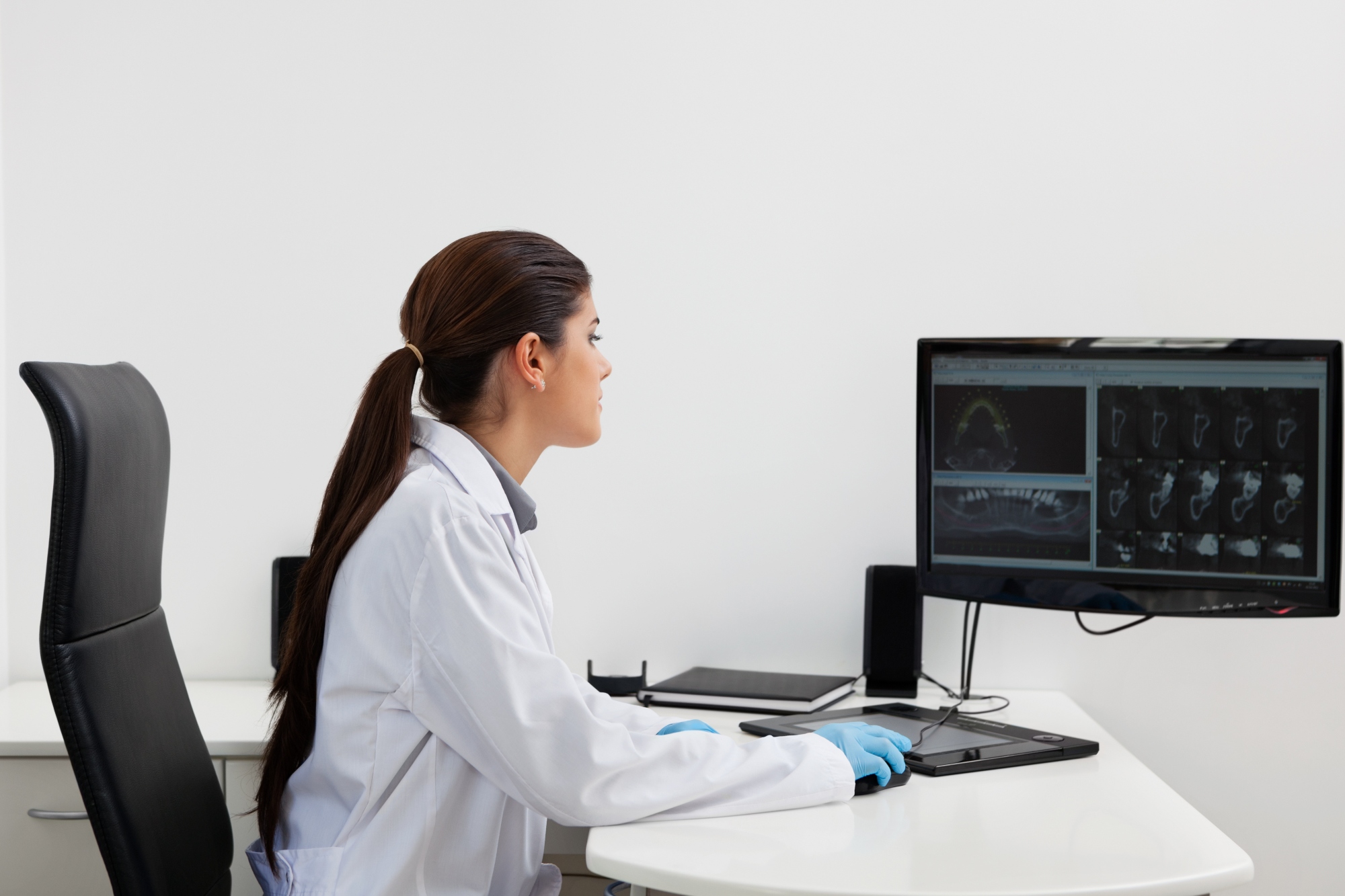 A dental professional in a white lab coat and blue gloves sits at a desk, viewing dental X-ray and CT images on a computer monitor.