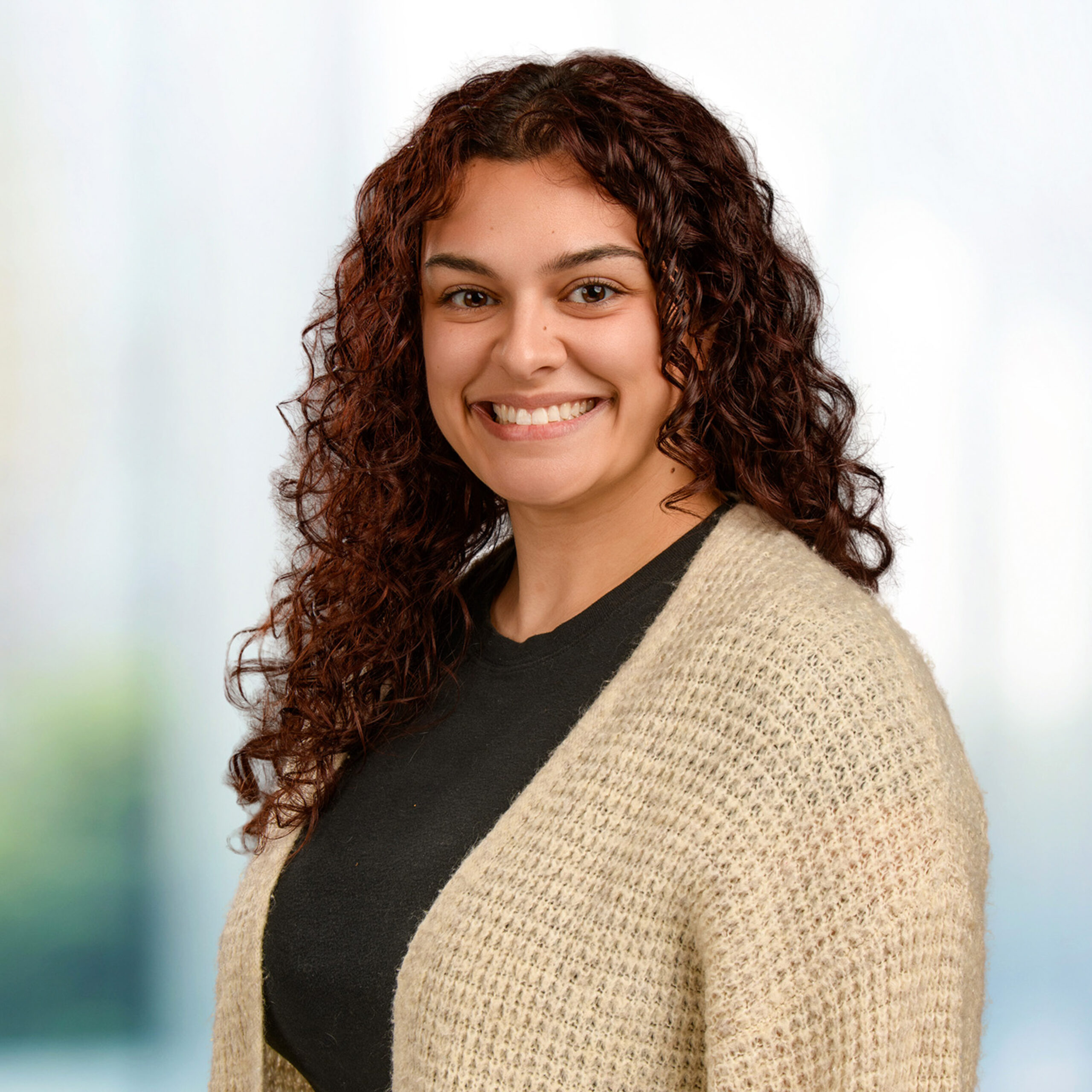 Smiling woman with curly dark brown hair wearing a beige knit cardigan over a black top against a blurred background