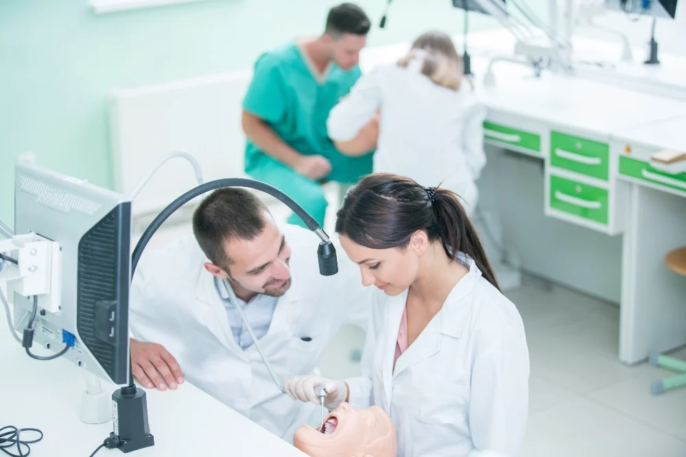 A female dental student in a white coat works on a dental mannequin with a dental tool while a male instructor watches over her in a bright training clinic with green-trimmed cabinets and another pair practicing in the background.