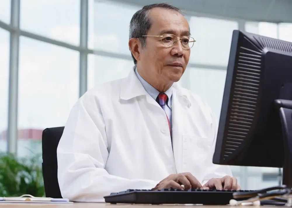 Older male doctor wearing a white lab coat and tie types on a keyboard while looking at a desktop monitor in a bright office with large curved windows.
