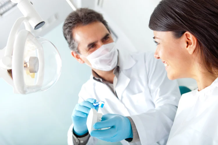 Dentist in a white coat and surgical mask, wearing blue gloves, demonstrating brushing technique on a dental model to a smiling female patient in a bright dental exam room.