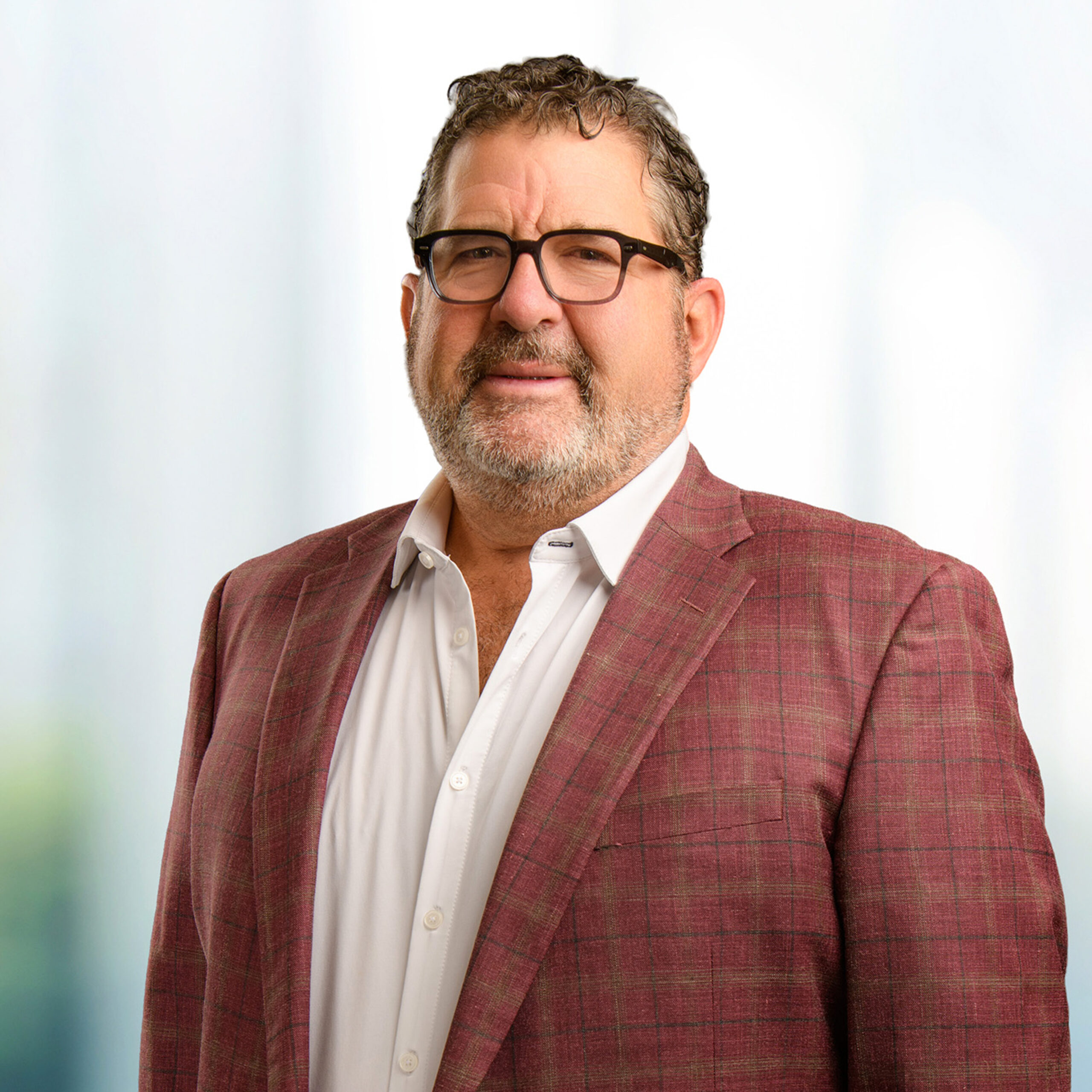 Middle-aged man with glasses and curly hair, wearing a white shirt and red checked blazer against a blurred background.