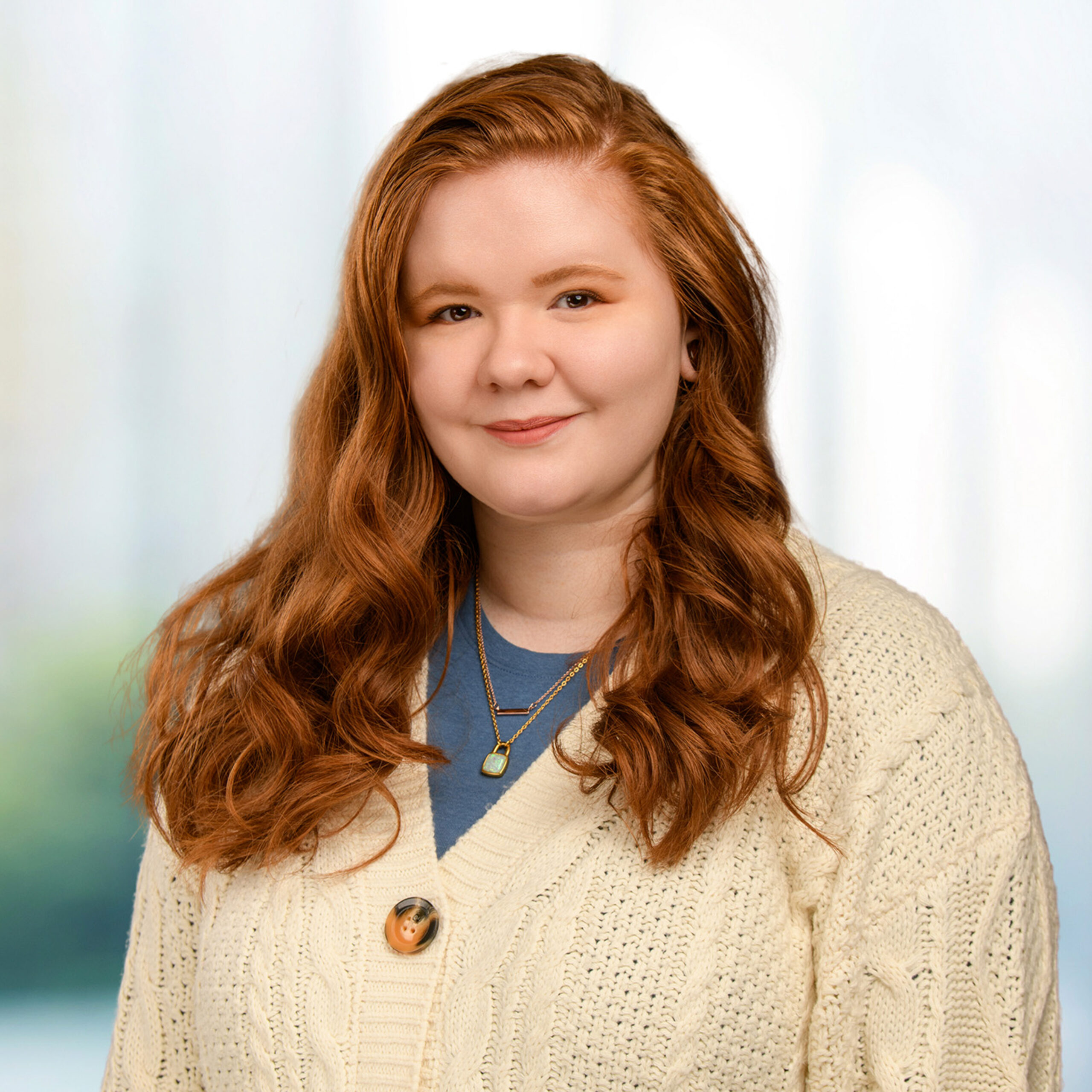 Young woman with long wavy red hair wearing a cream cable-knit cardigan and layered pendant necklaces, smiling.