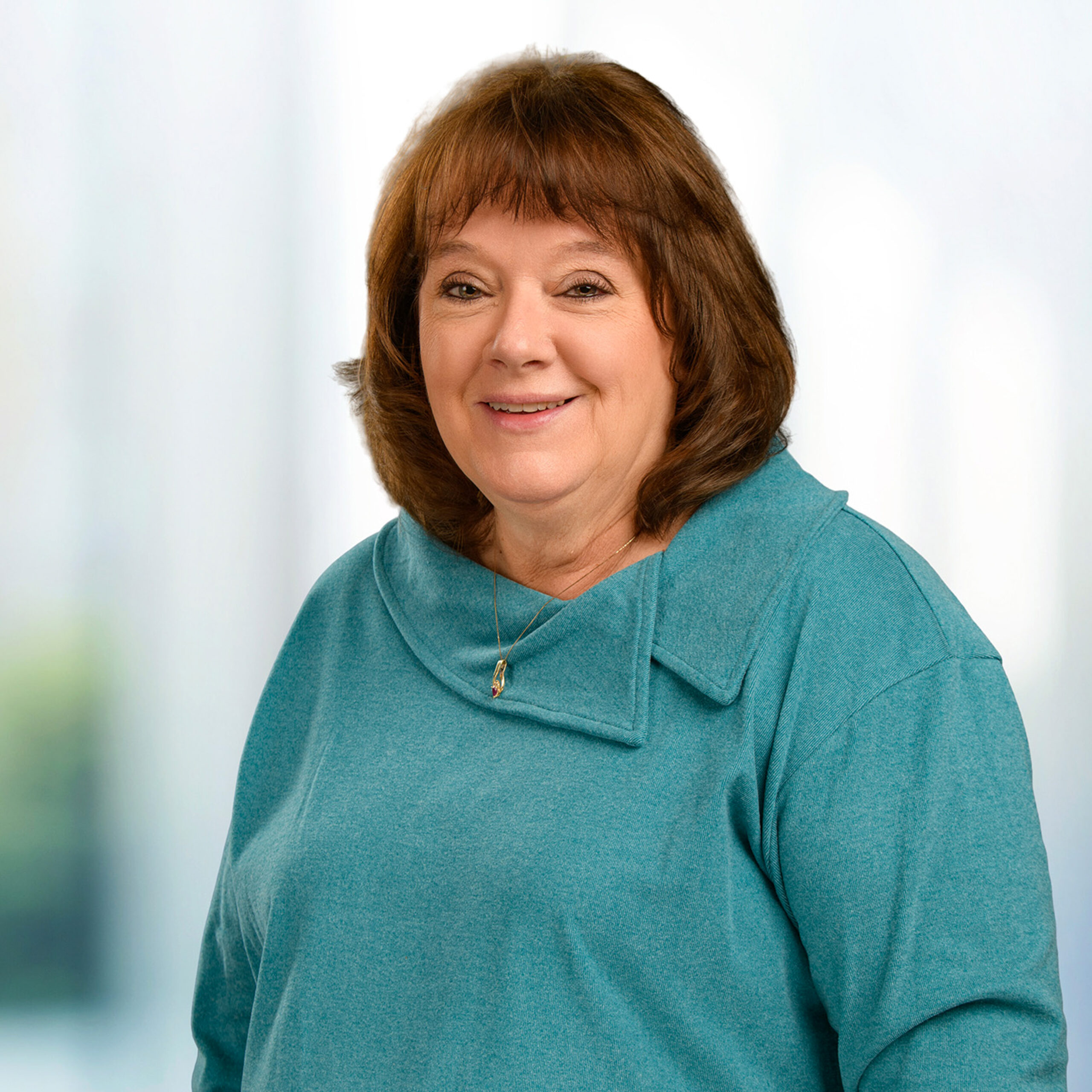 Smiling middle-aged woman with brown shoulder-length hair and bangs, wearing a teal cowl-neck top and small pendant necklace against a soft blurred background.