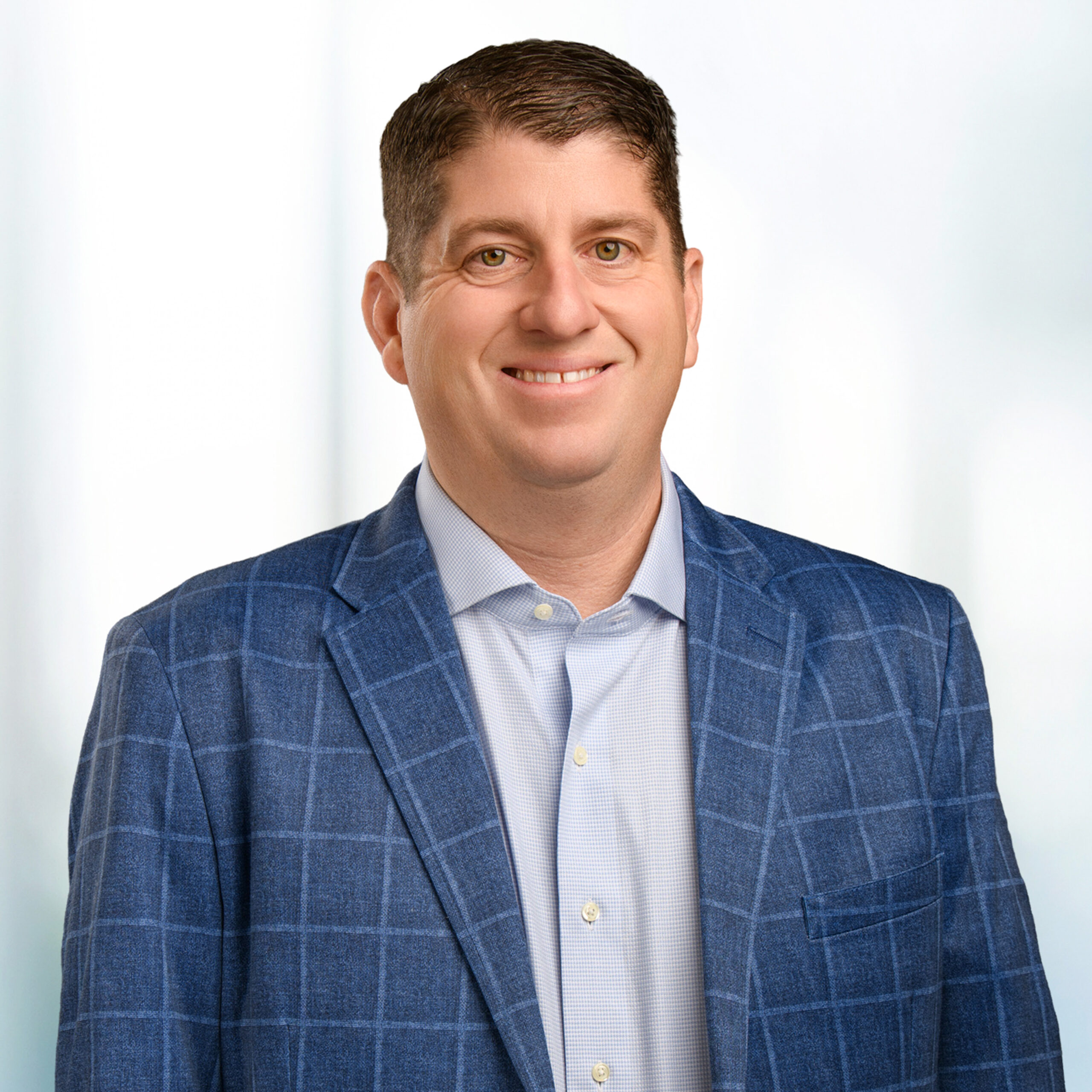 Smiling middle-aged man in a blue checked blazer and light dress shirt against a soft, bright background.