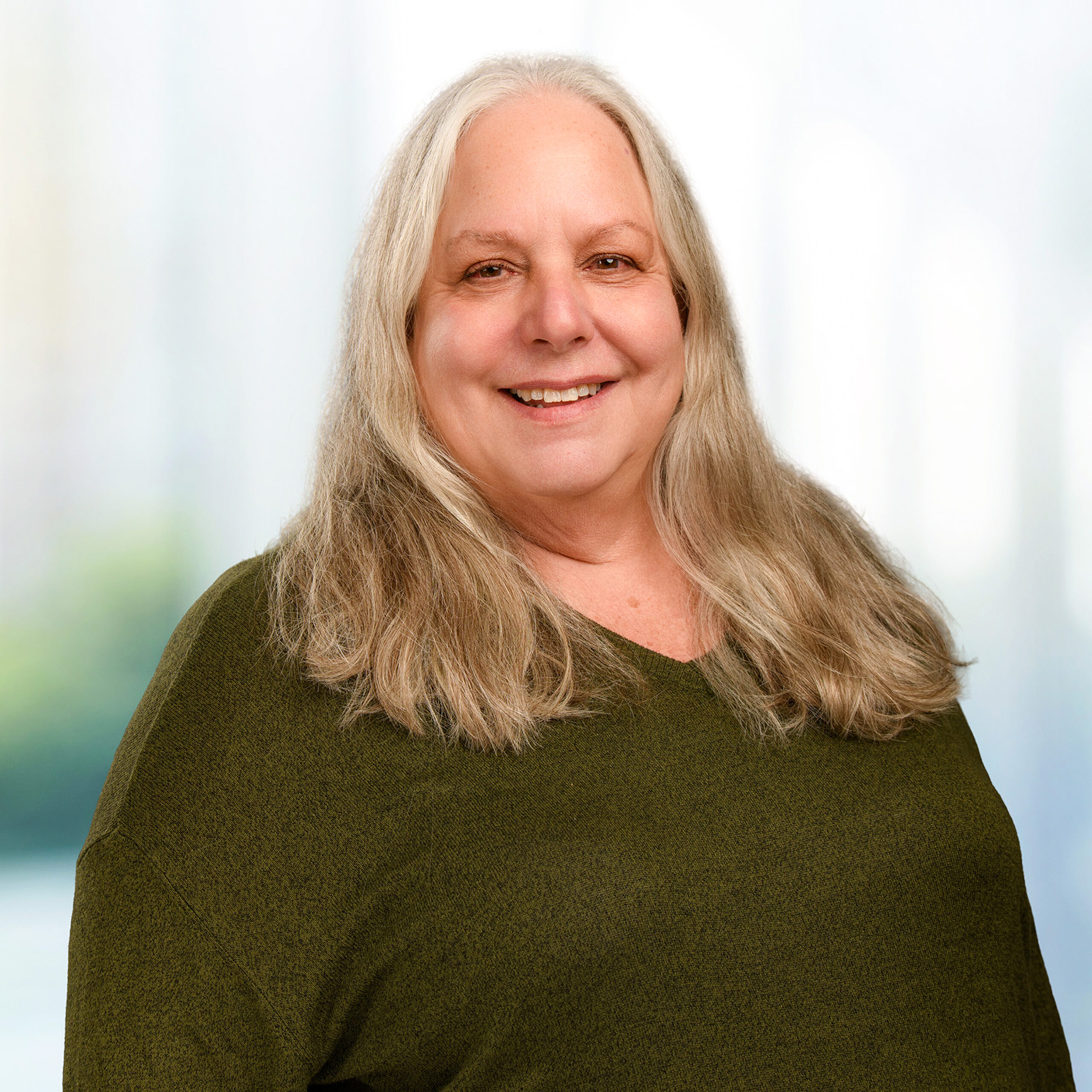 Smiling older woman with long gray hair wearing an olive-green sweater against a soft, blurred background.
