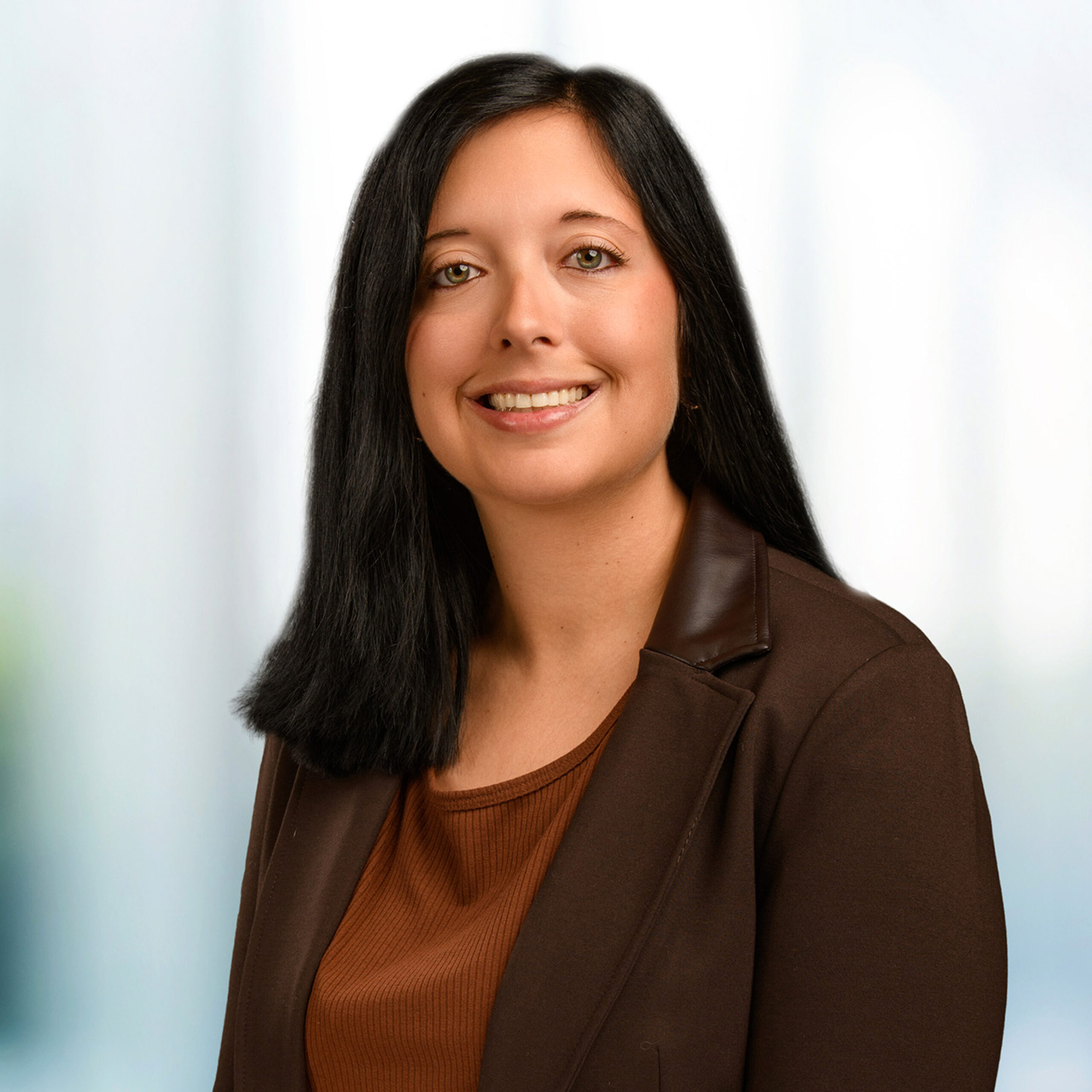 Smiling woman with long straight black hair wearing a brown blazer and rust-colored top in a light studio portrait.