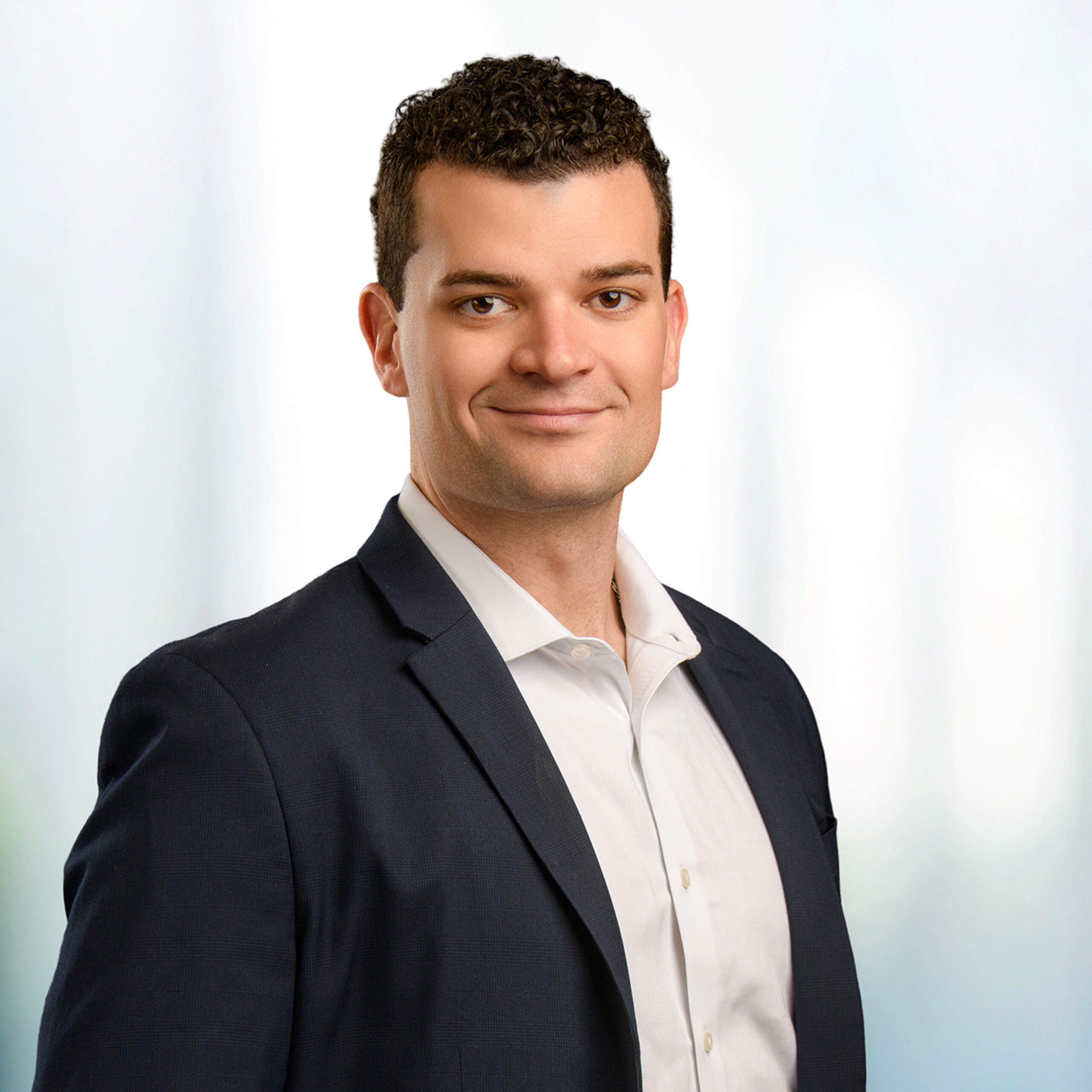 Smiling man with short curly dark hair wearing a dark blazer and white dress shirt, posed against a soft light blurred background.