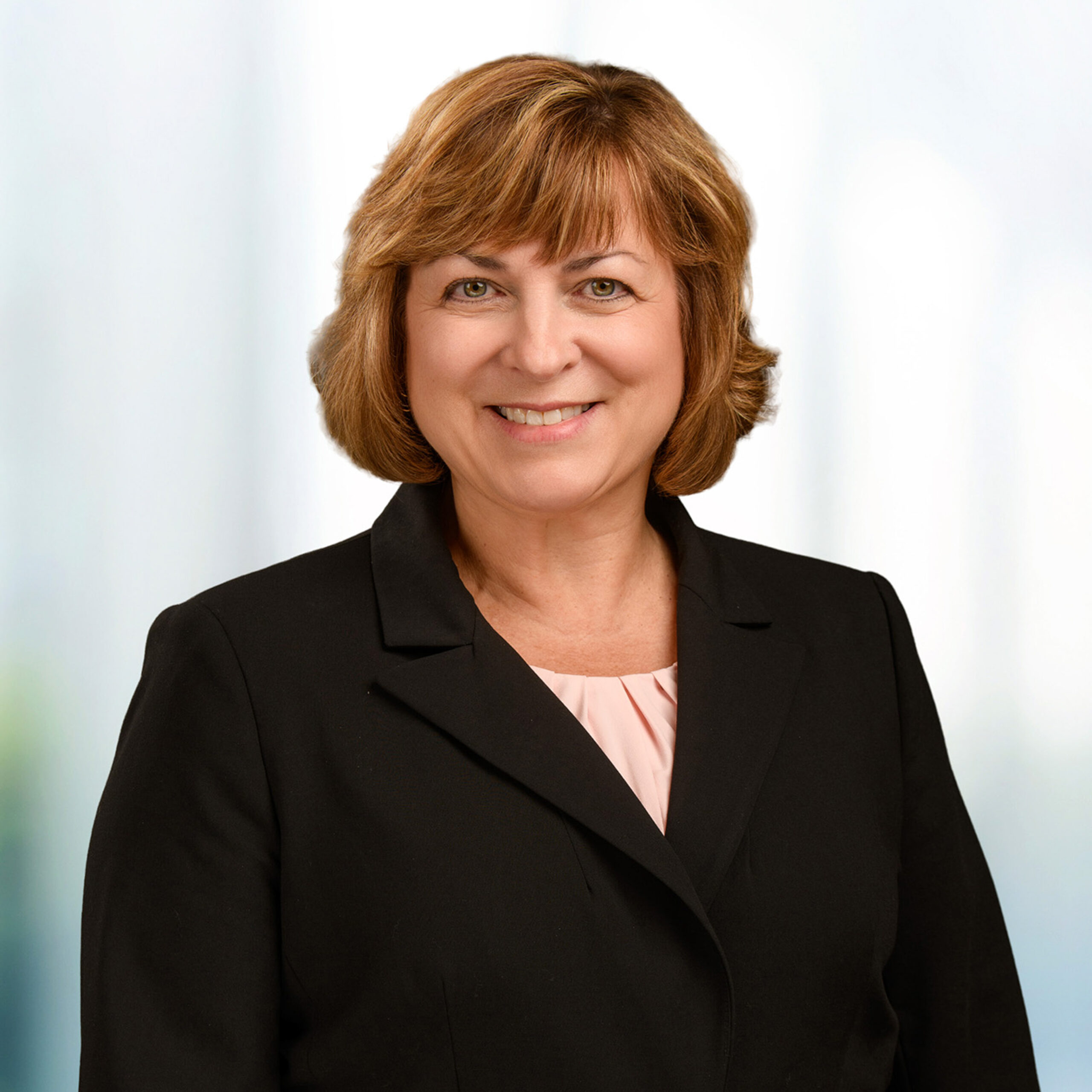 Woman with light brown hair smiling in a black blazer and pale pink blouse against a soft blurred background.