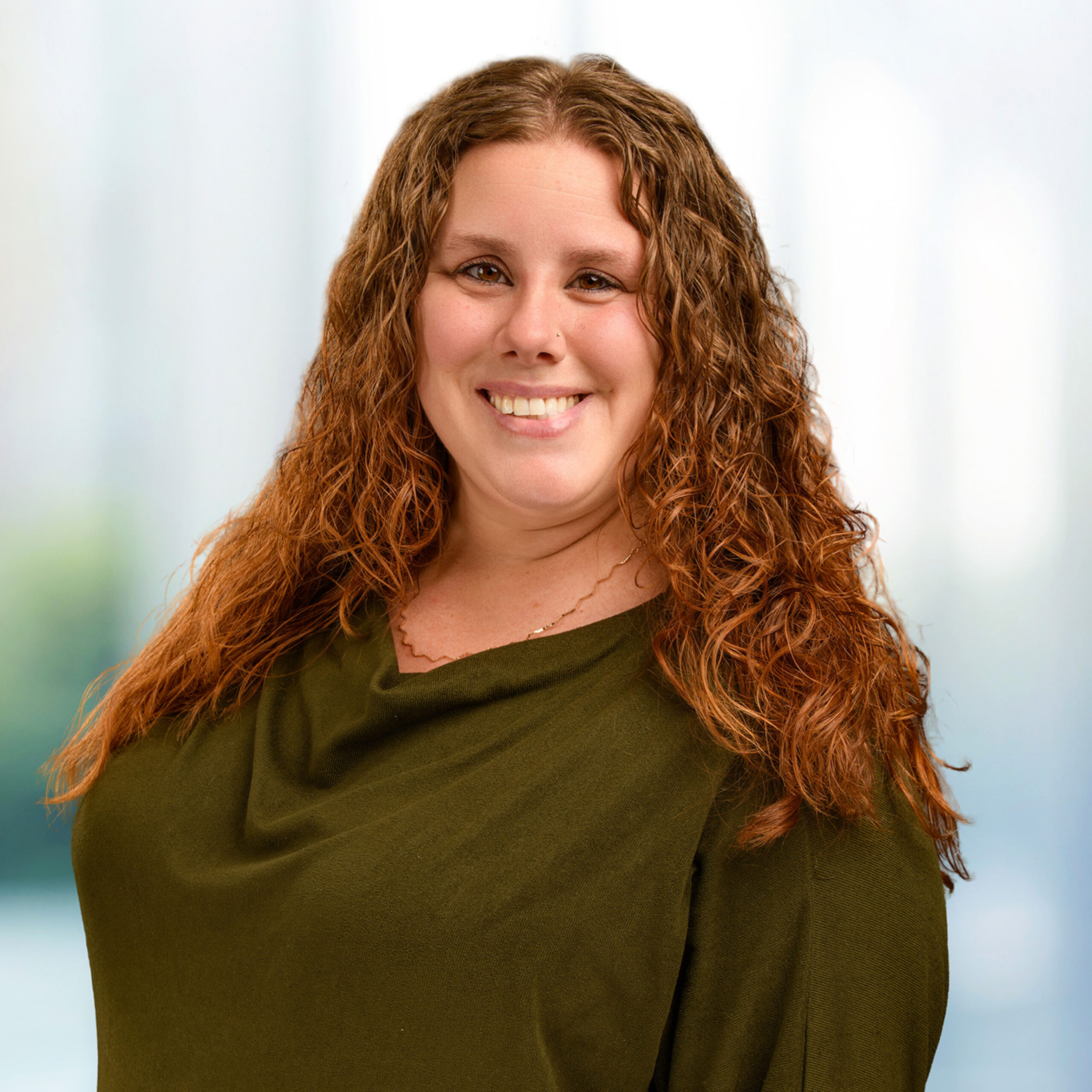 Smiling woman with long curly reddish-brown hair wearing an olive-green top against a soft, blurred background