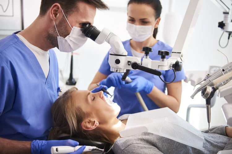 Male dentist peers through a dental microscope while a masked assistant holds suction and instruments as they treat a reclining female patient wearing a paper bib in a bright dental operatory.
