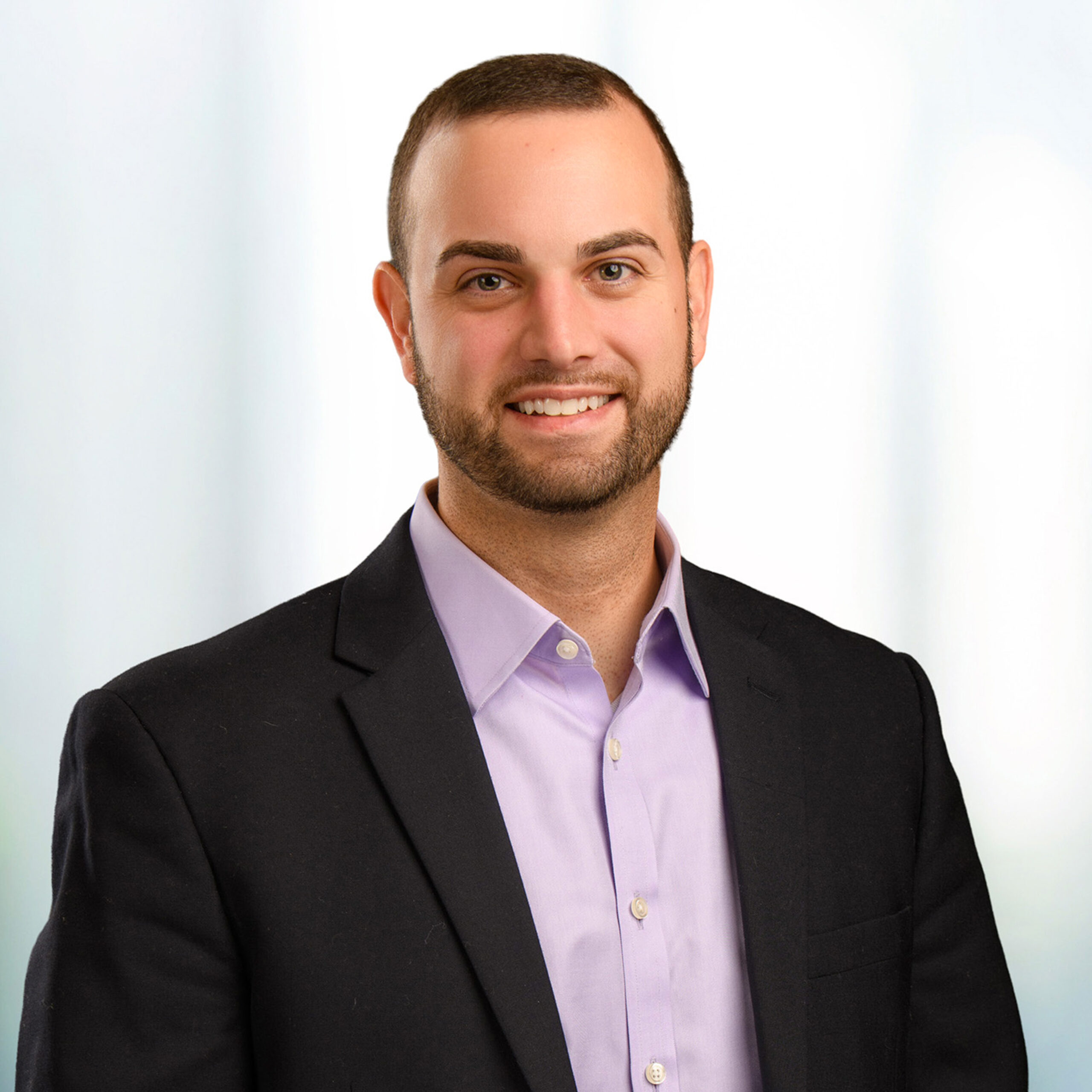 Smiling man with short hair and trimmed beard wearing a dark blazer and lavender shirt against a light blurred background.