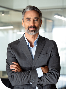 Middle-aged man with salt-and-pepper hair and beard wearing a dark gray suit and light blue shirt, standing with arms crossed in a modern glass-walled office and looking toward the camera.