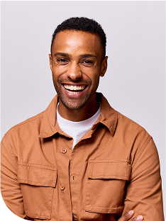 Smiling young adult with short curly hair wearing a light brown button-up jacket over a white T-shirt against a plain light gray background.