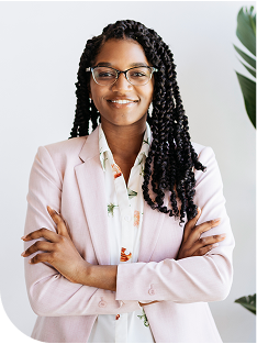 Confident woman with long braided hair and glasses, smiling with arms crossed while wearing a light pink blazer over a patterned blouse against a bright neutral background with a tall green plant.
