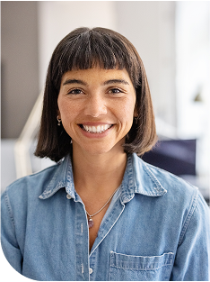 Smiling person with short dark hair and blunt bangs, wearing a denim button-up shirt and delicate necklaces, seated in a bright modern office setting.