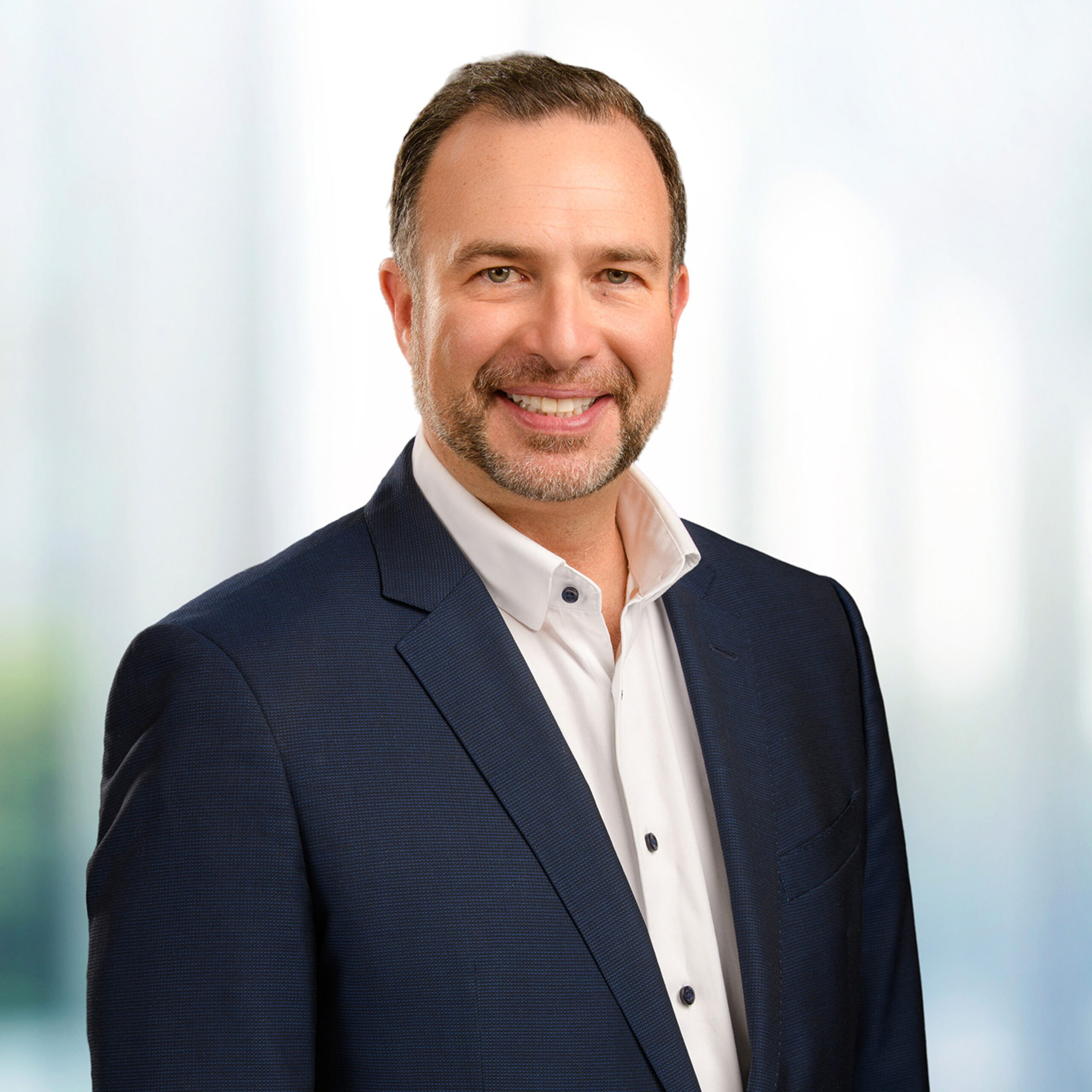 Smiling man with short dark hair and trimmed beard in a white shirt and navy suit against a bright blurred background.