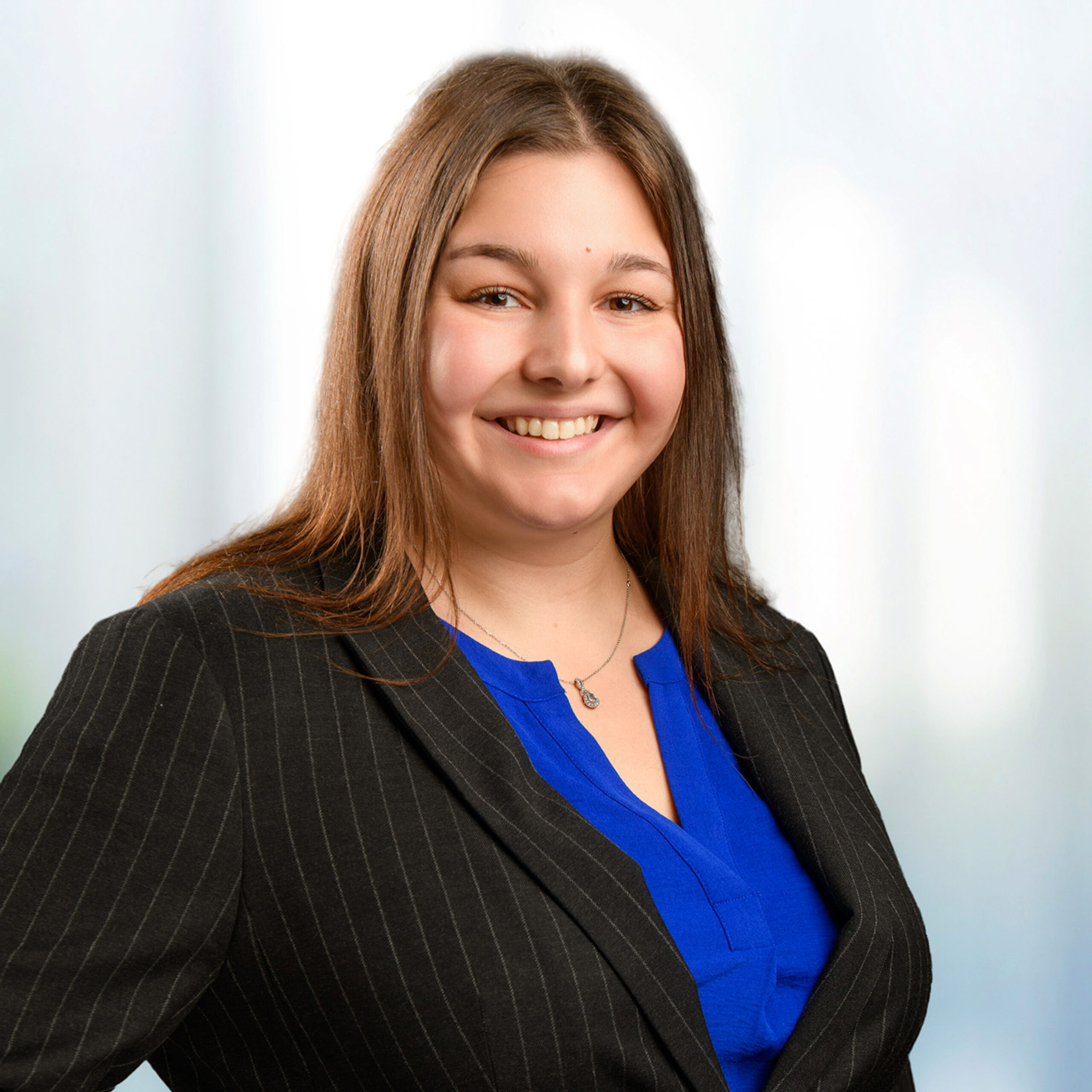 Smiling woman with brown hair wearing a pinstripe blazer, blue blouse, and small pendant against a soft light background.