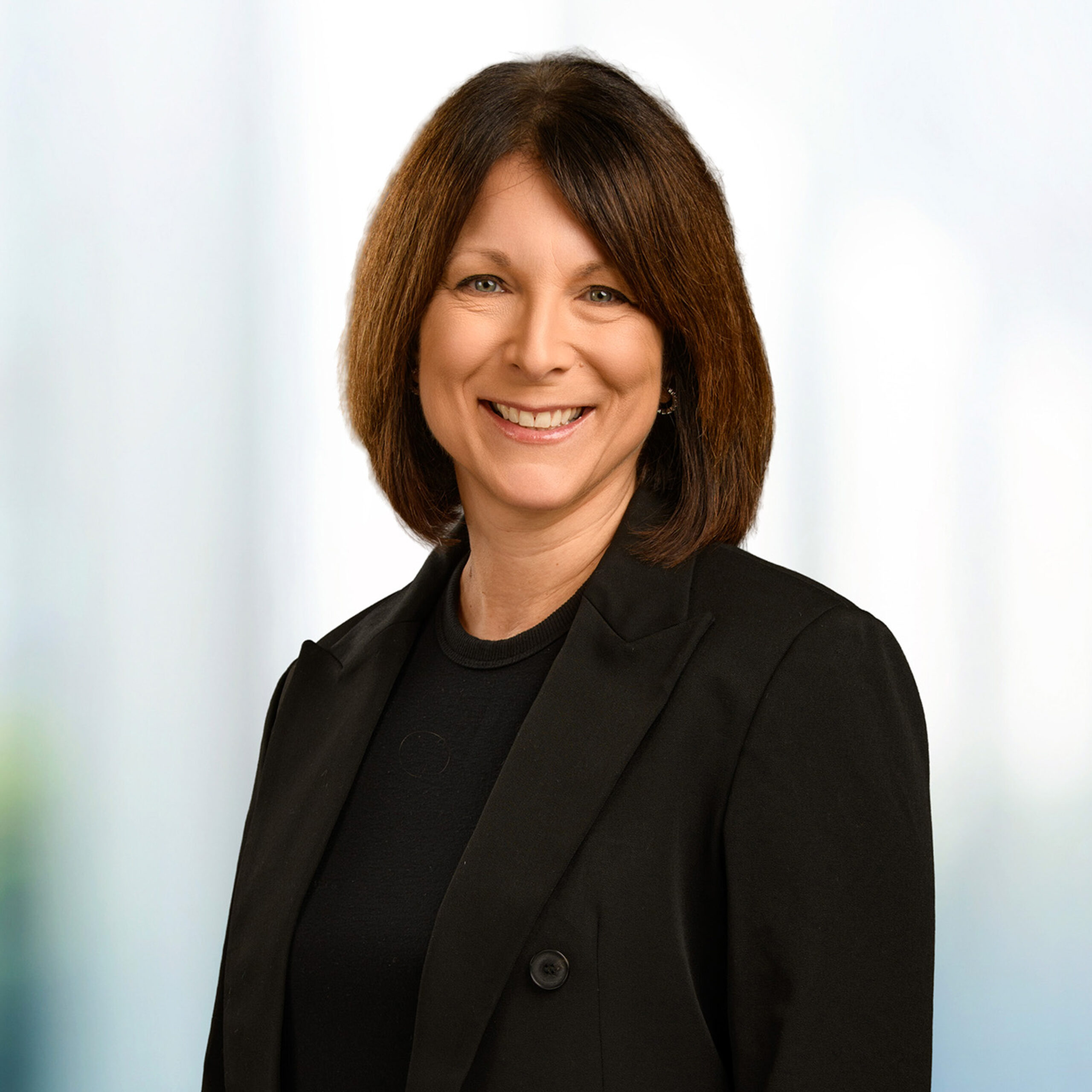 Smiling woman with shoulder-length brown hair wearing a black blazer against a soft, light blurred background
