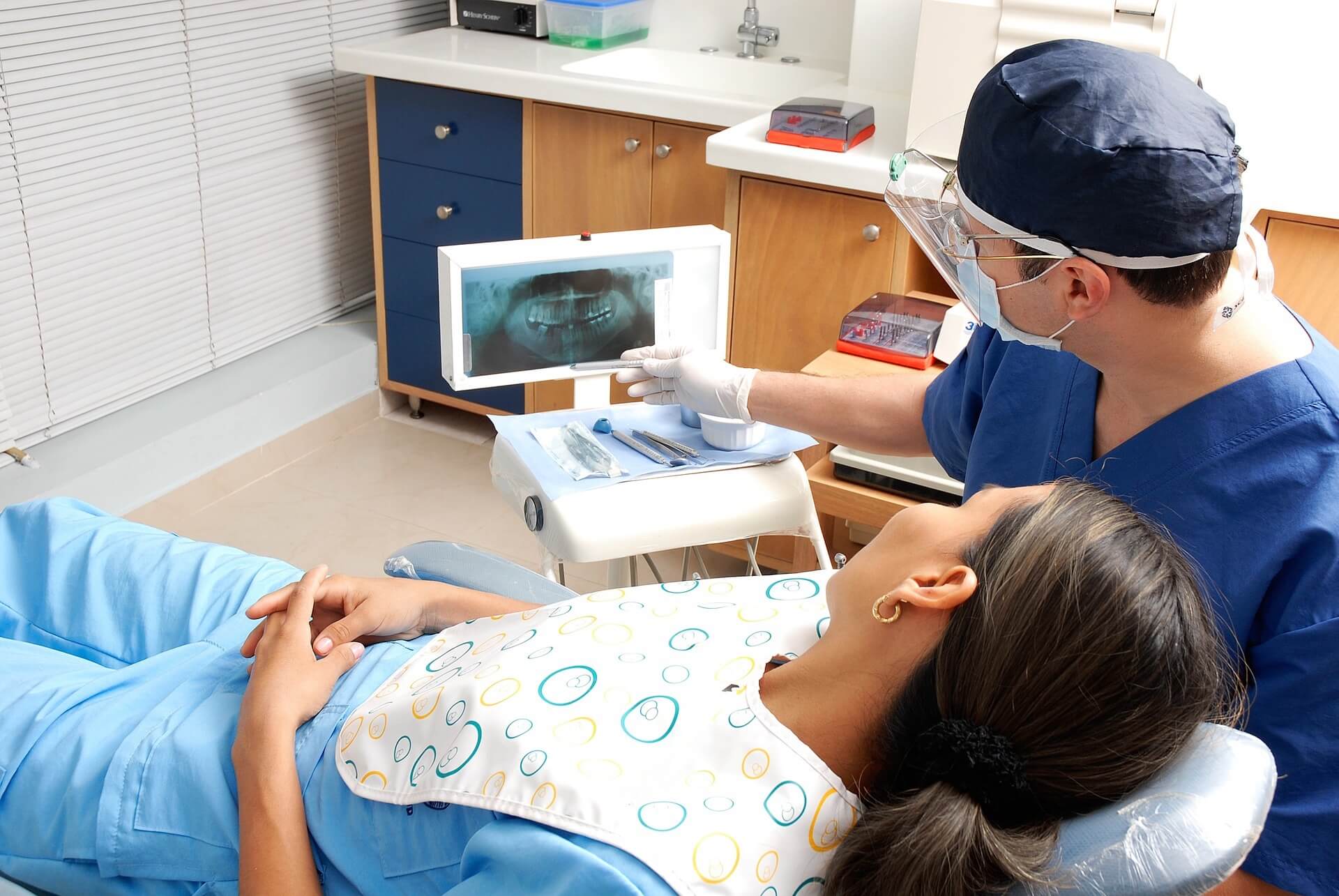 Dentist in blue scrubs, mask and face shield points at a dental X‑ray on a lightbox while a patient reclines in the chair wearing a patterned bib and folded hands, with a tray of dental instruments nearby.