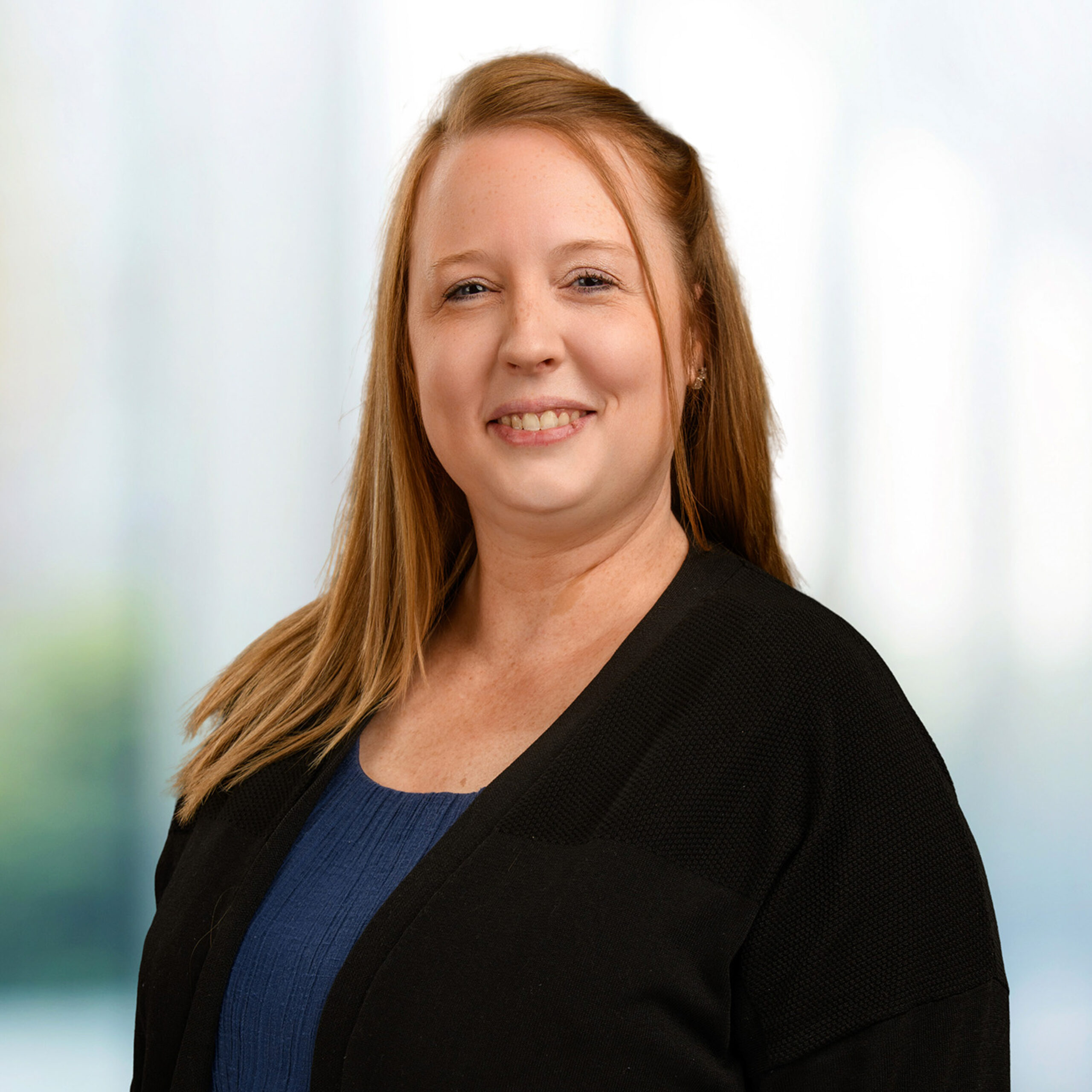 Smiling woman with long light brown hair wearing a blue top and black cardigan against a soft blurred background.